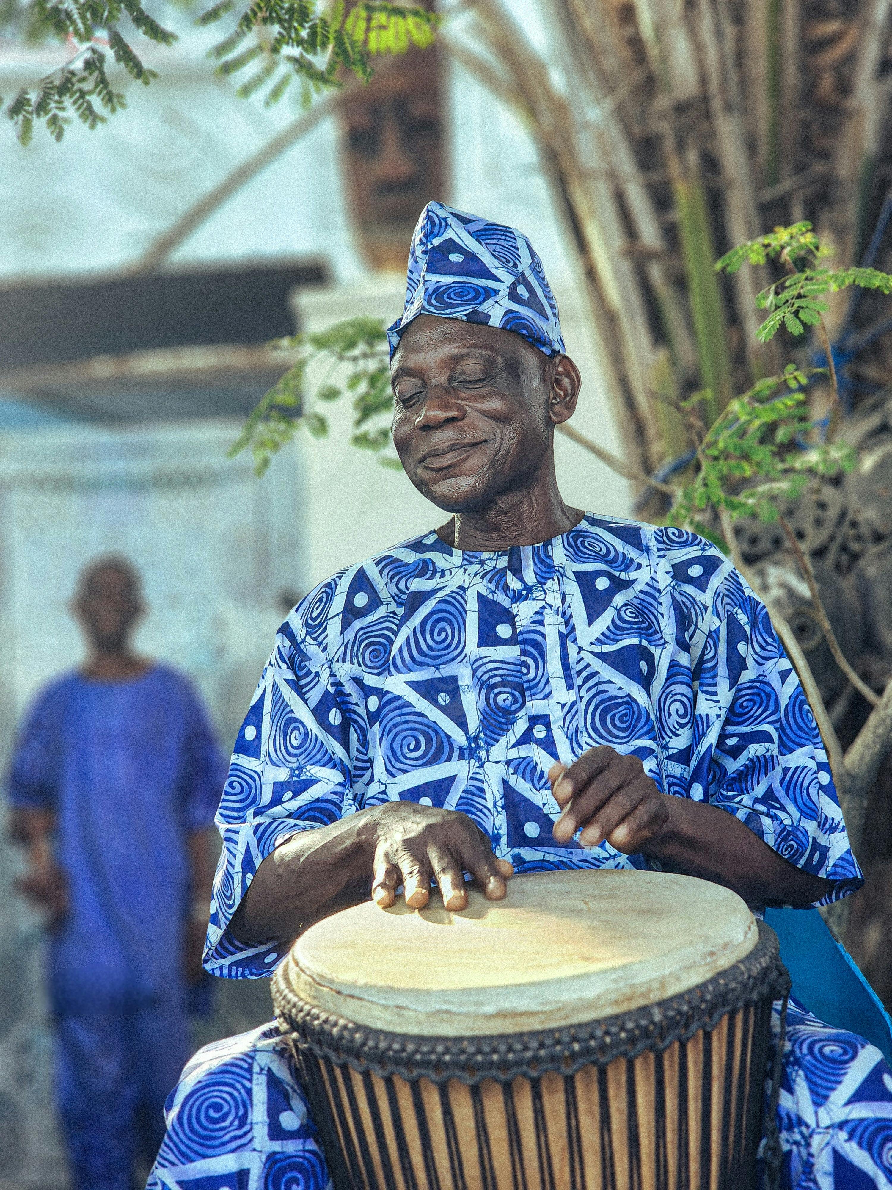 Elderly man joyfully playing African drum in vibrant traditional attire outdoors.