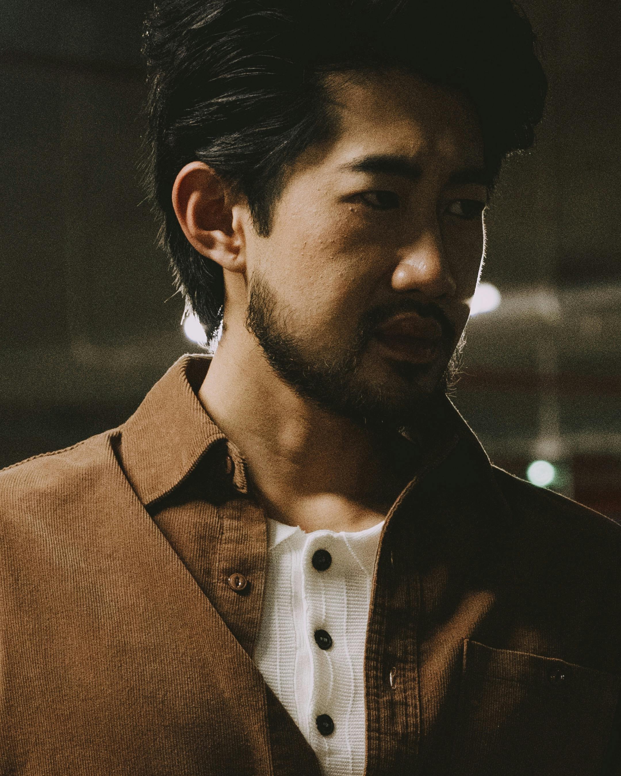 A contemplative man in a brown jacket stands in low light, creating a moody atmosphere.
