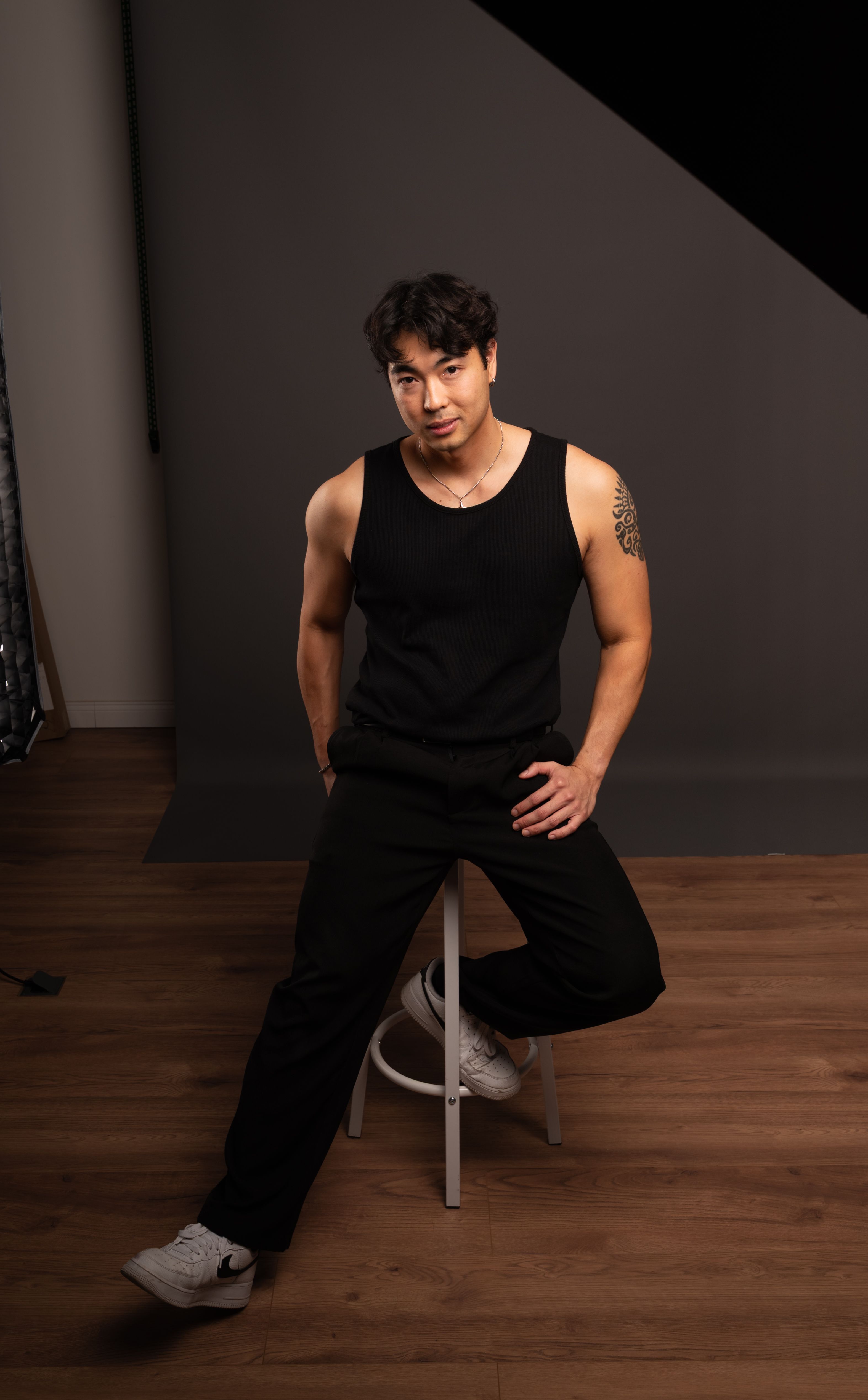 Portrait of a young man in a black tank top seated on a stool in a studio setting.