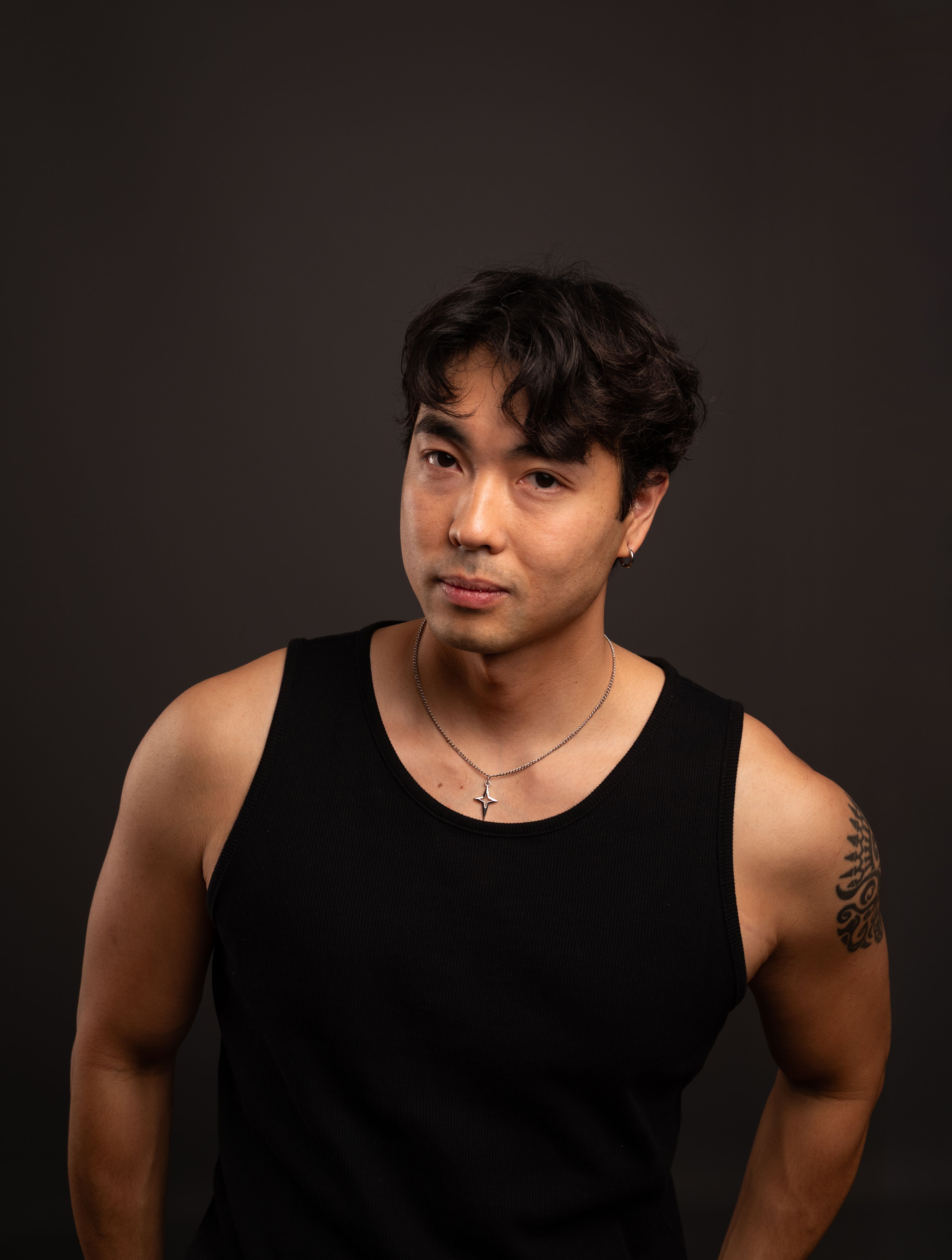 Studio portrait of a man in a black tank top with a tattoo on his arm, dark background.