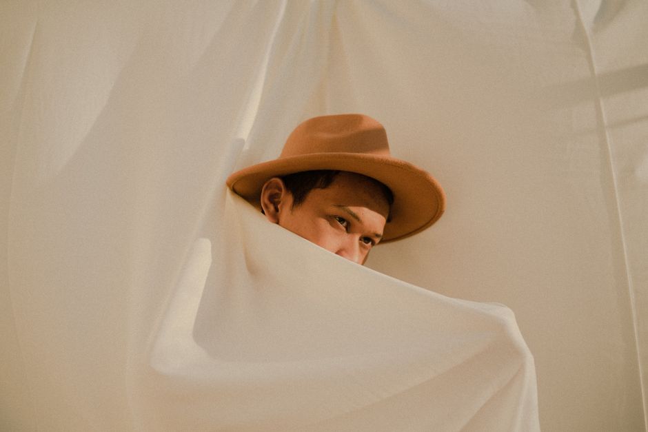 A young man in a hat peeks from behind white curtains in a soft light setting.