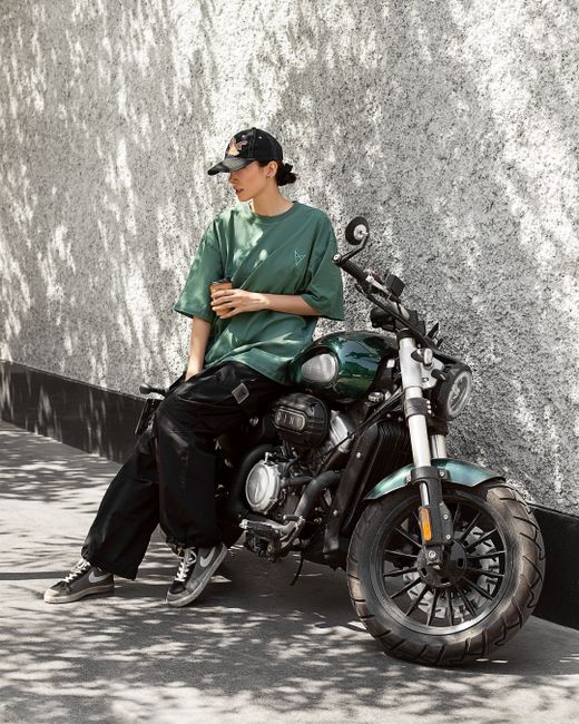 Woman in casual outfit resting on a motorcycle against a wall outside during the day.