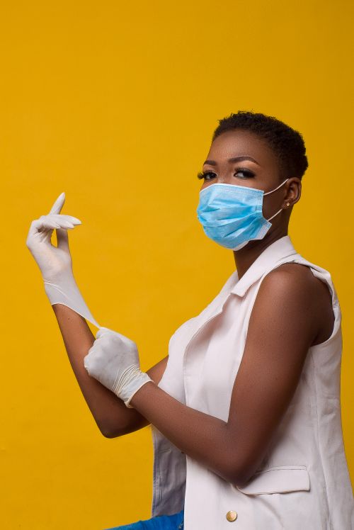 Free African woman posing with mask and gloves against a yellow backdrop, highlighting safety. Stock Photo