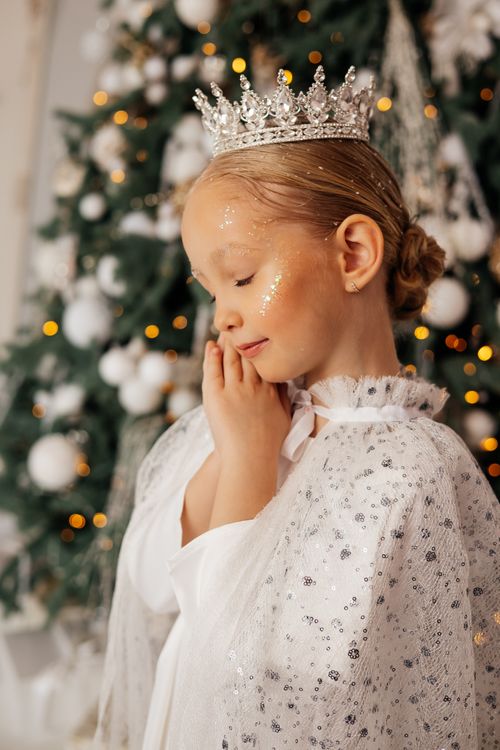 Free A young girl with a glittery crown in festive attire stands beside a decorated Christmas tree. Stock Photo
