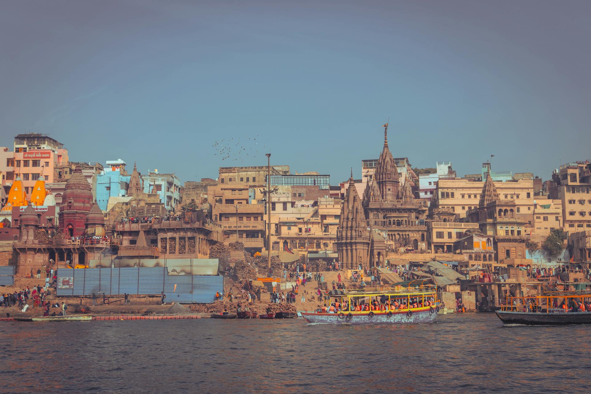 A vibrant view of Varanasi ghats lined with historic temples and busy riverbanks under a clear blue sky.