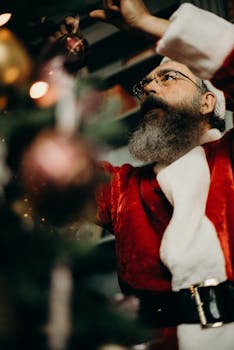 一位大胡子男人穿着圣诞老人服装，在室内用装饰品装饰圣诞树
