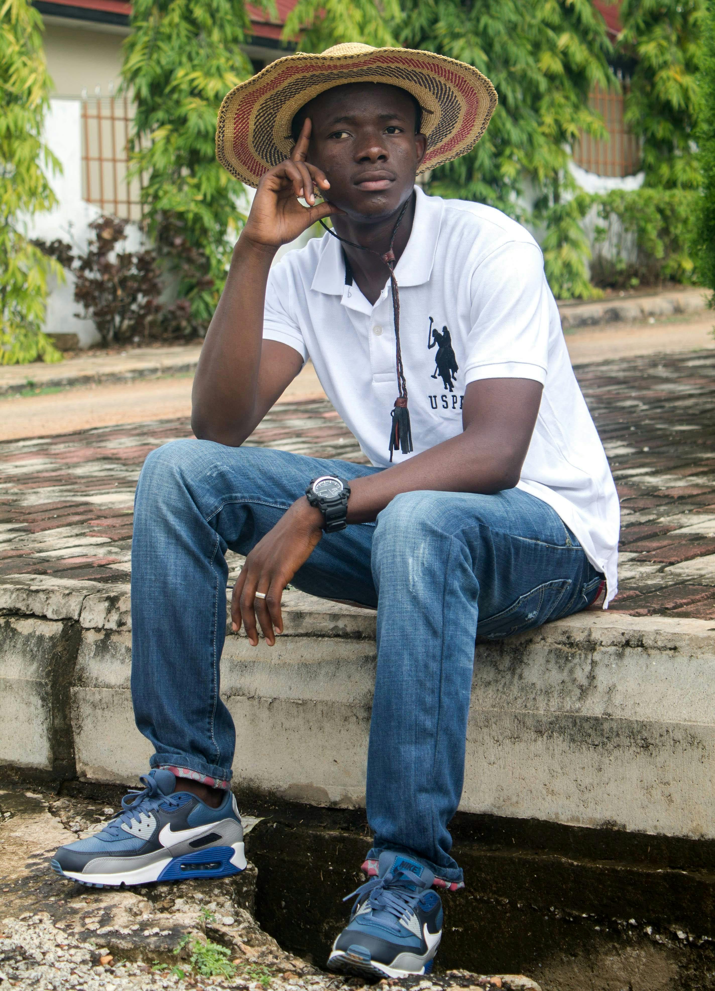 Casually dressed young man in straw hat sits on pavement edge, lost in thought outdoors.