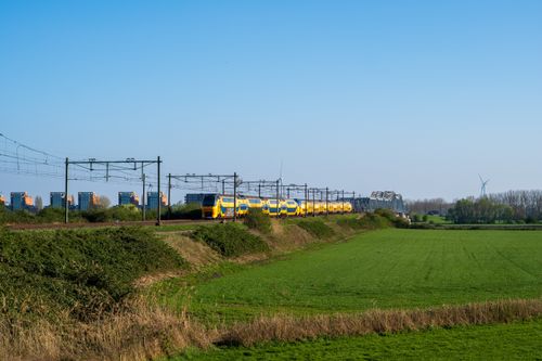 Free Yellow train crossing a rural area with green fields and distant city buildings under a clear blue sky. Stock Photo