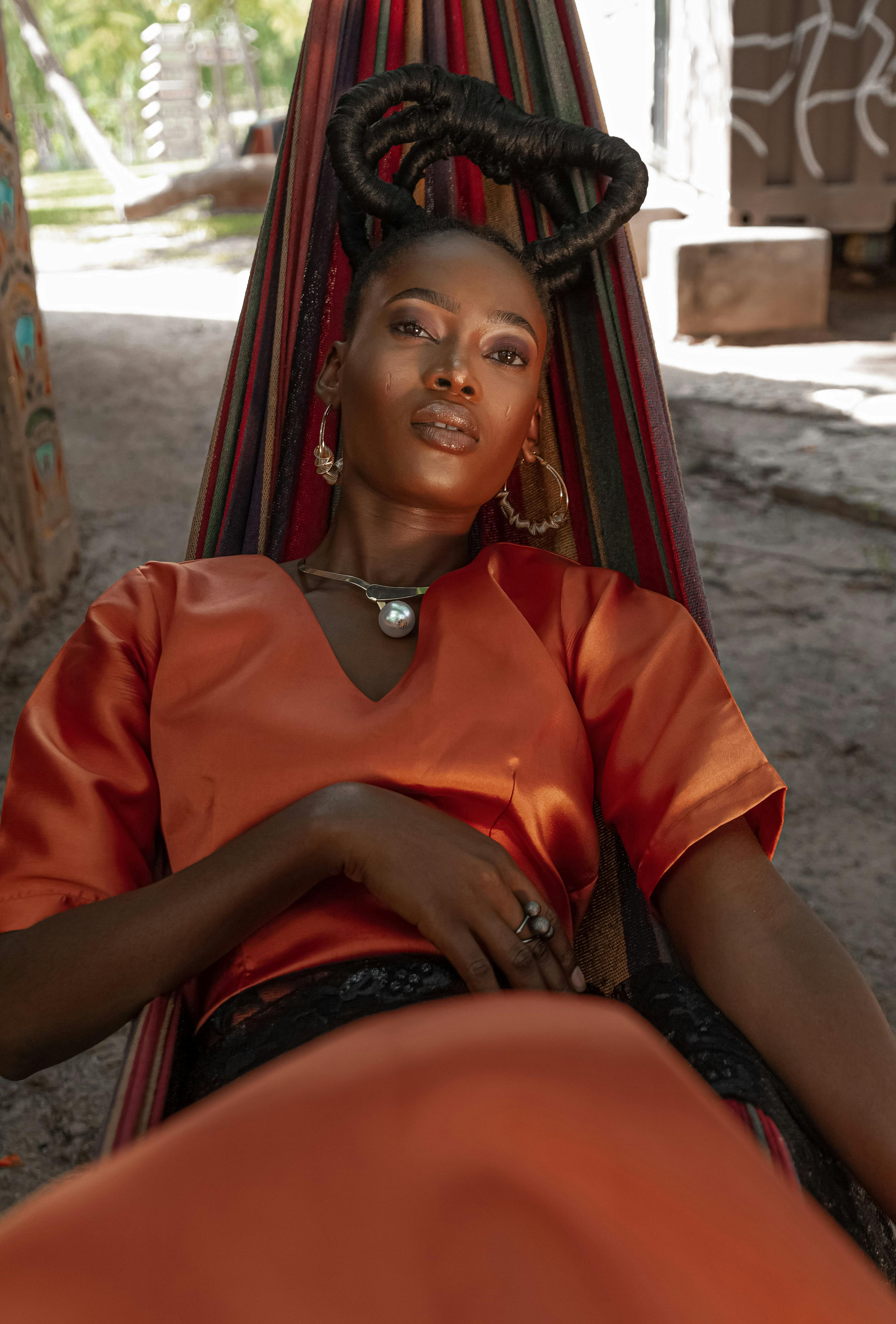 Stylish woman in an orange outfit reclines in a colorful hammock outdoors, exuding calm and elegance.