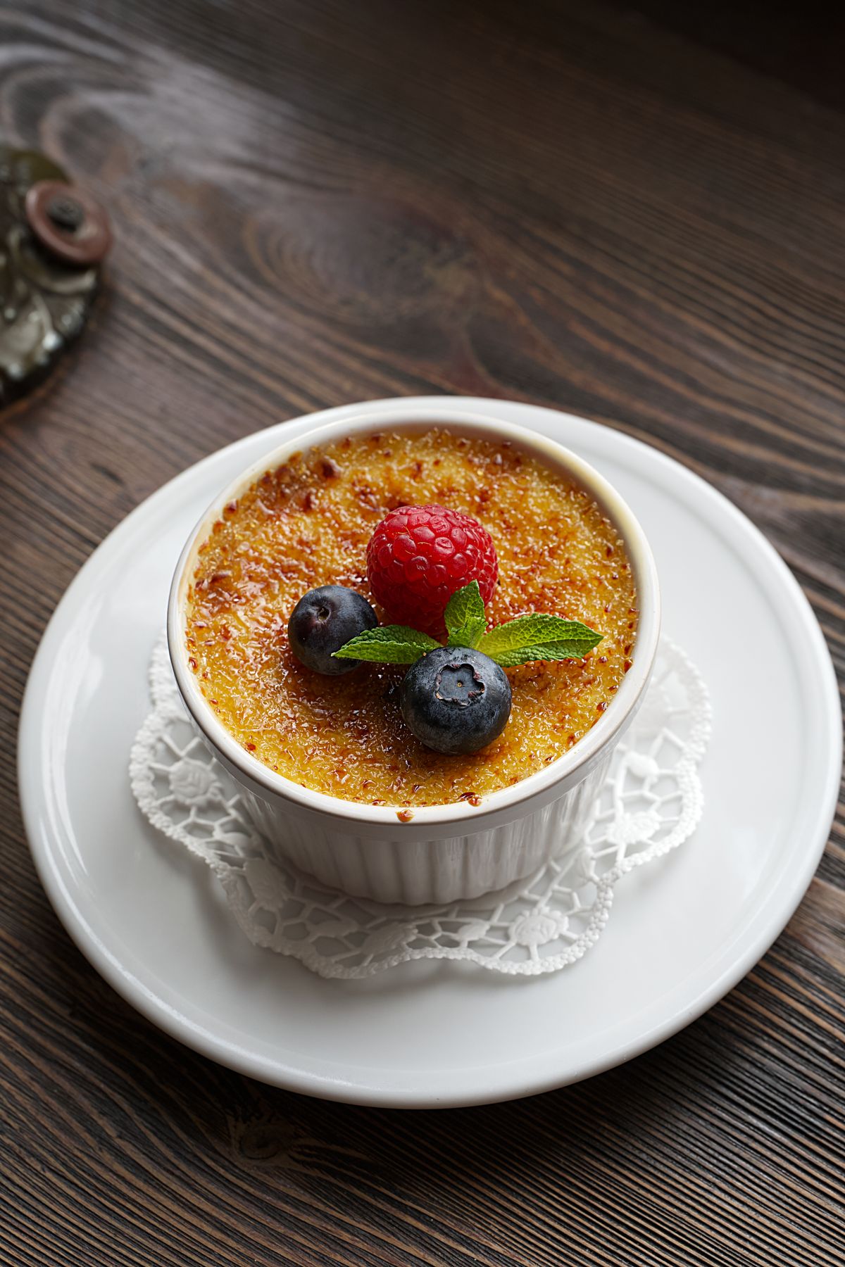 Creme brulee in fluted white ramekin topped with raspberry, blueberries and mint on doily plate