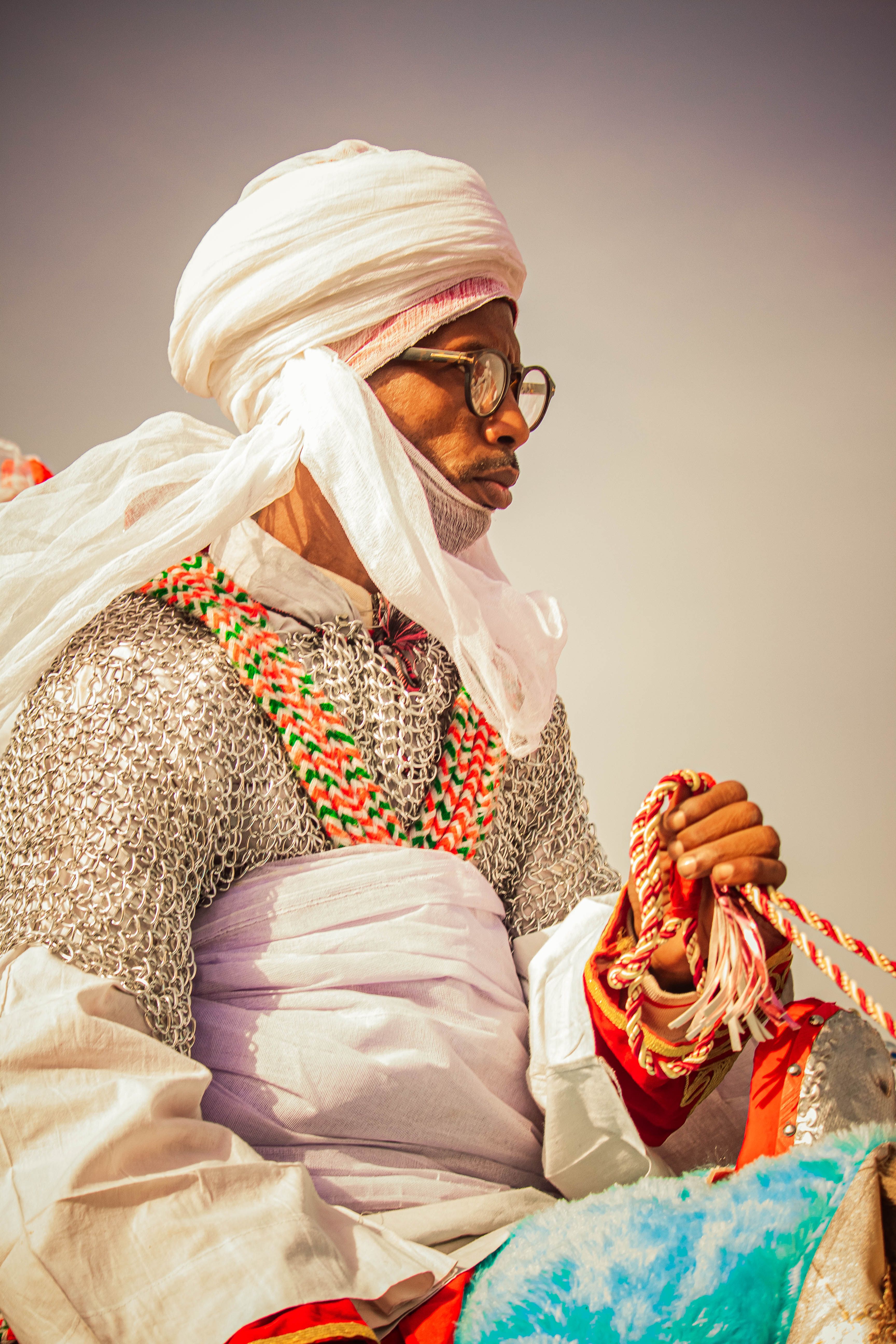 A man in traditional attire, with a white turban and chainmail, rides a decorated camel in a desert setting.