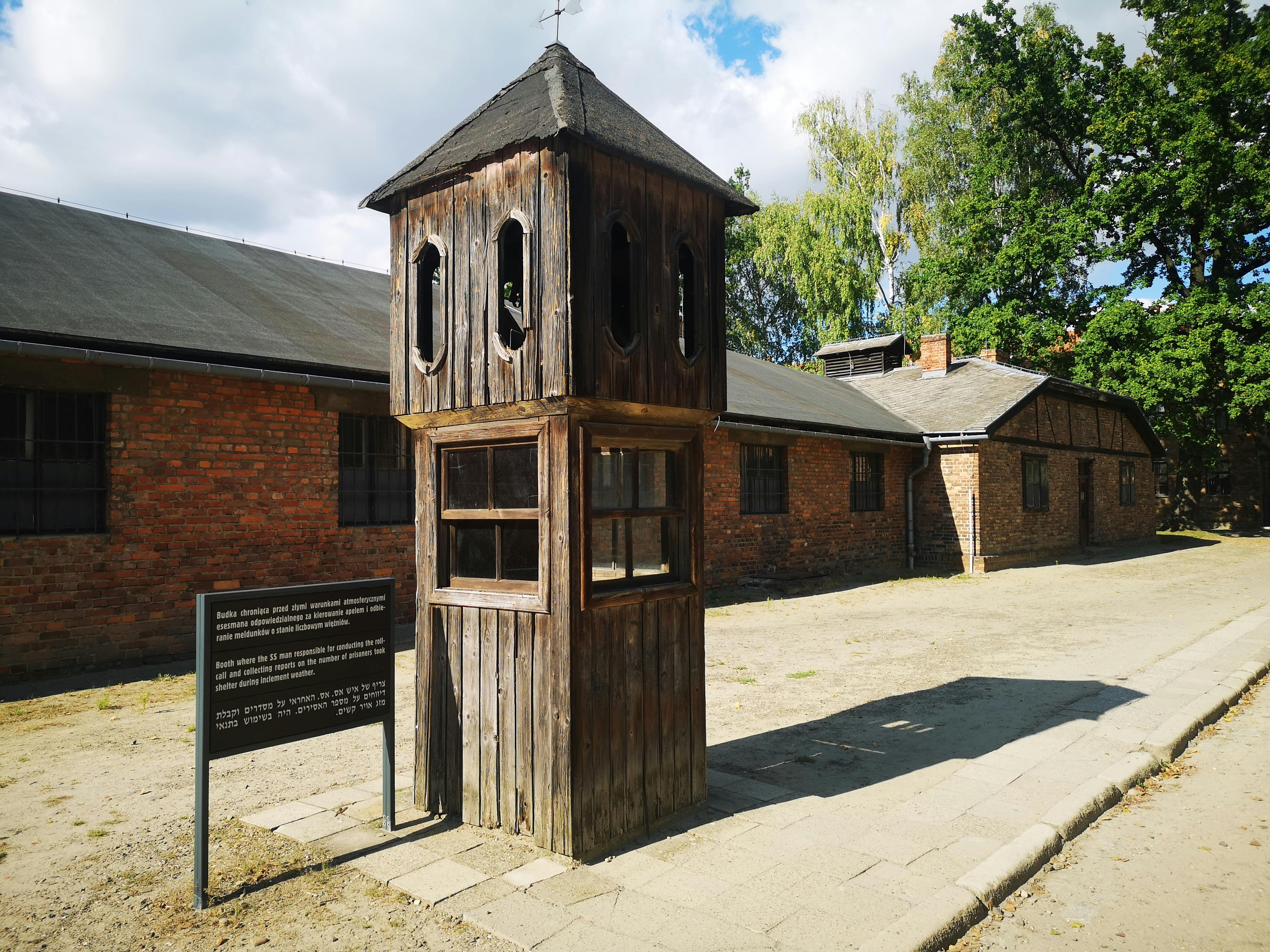 A wooden guard tower stands in front of brick buildings at a historic concentration camp site.