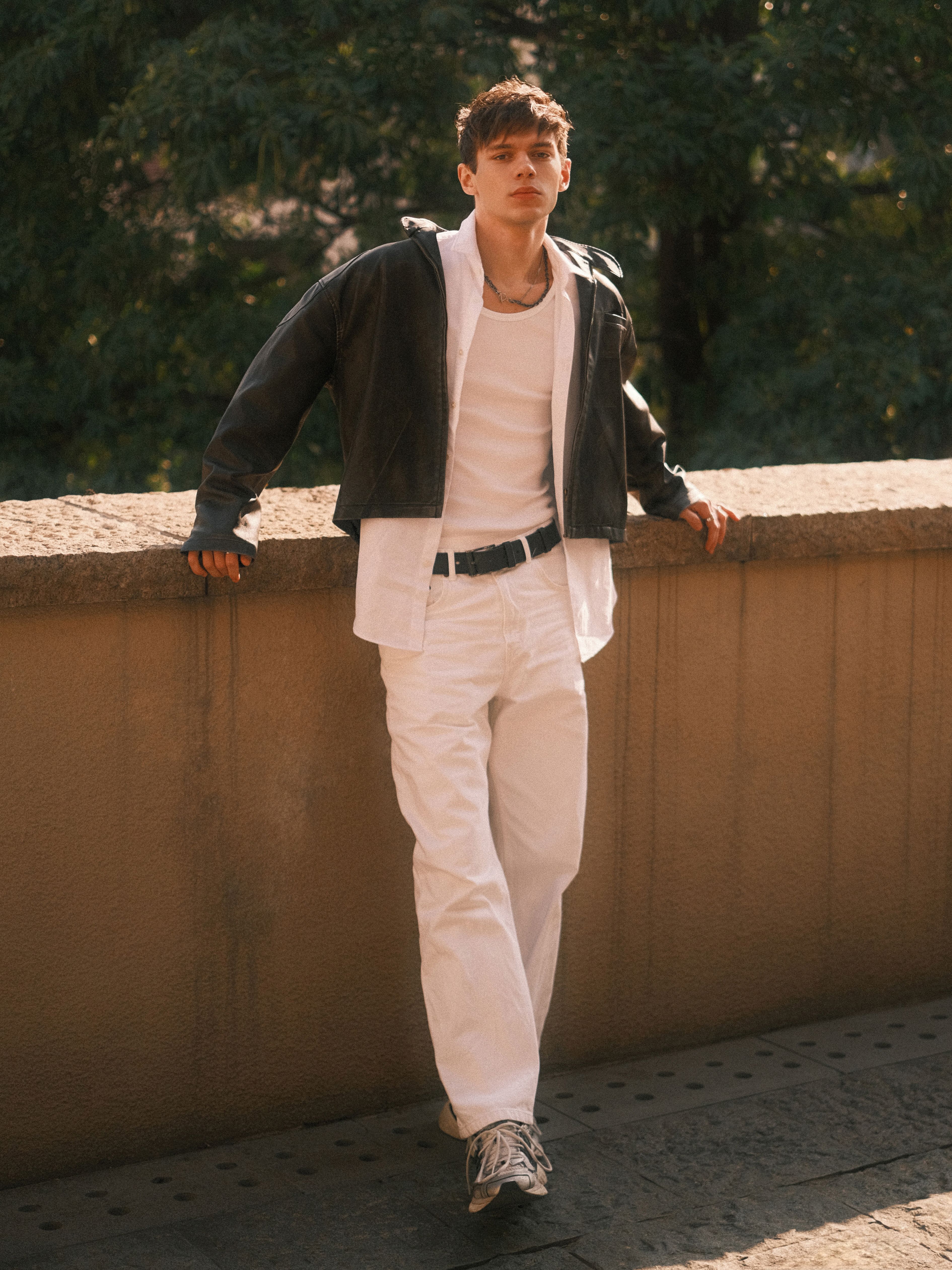 Young man in a leather jacket and casual wear standing outdoors by a stone wall in daylight.