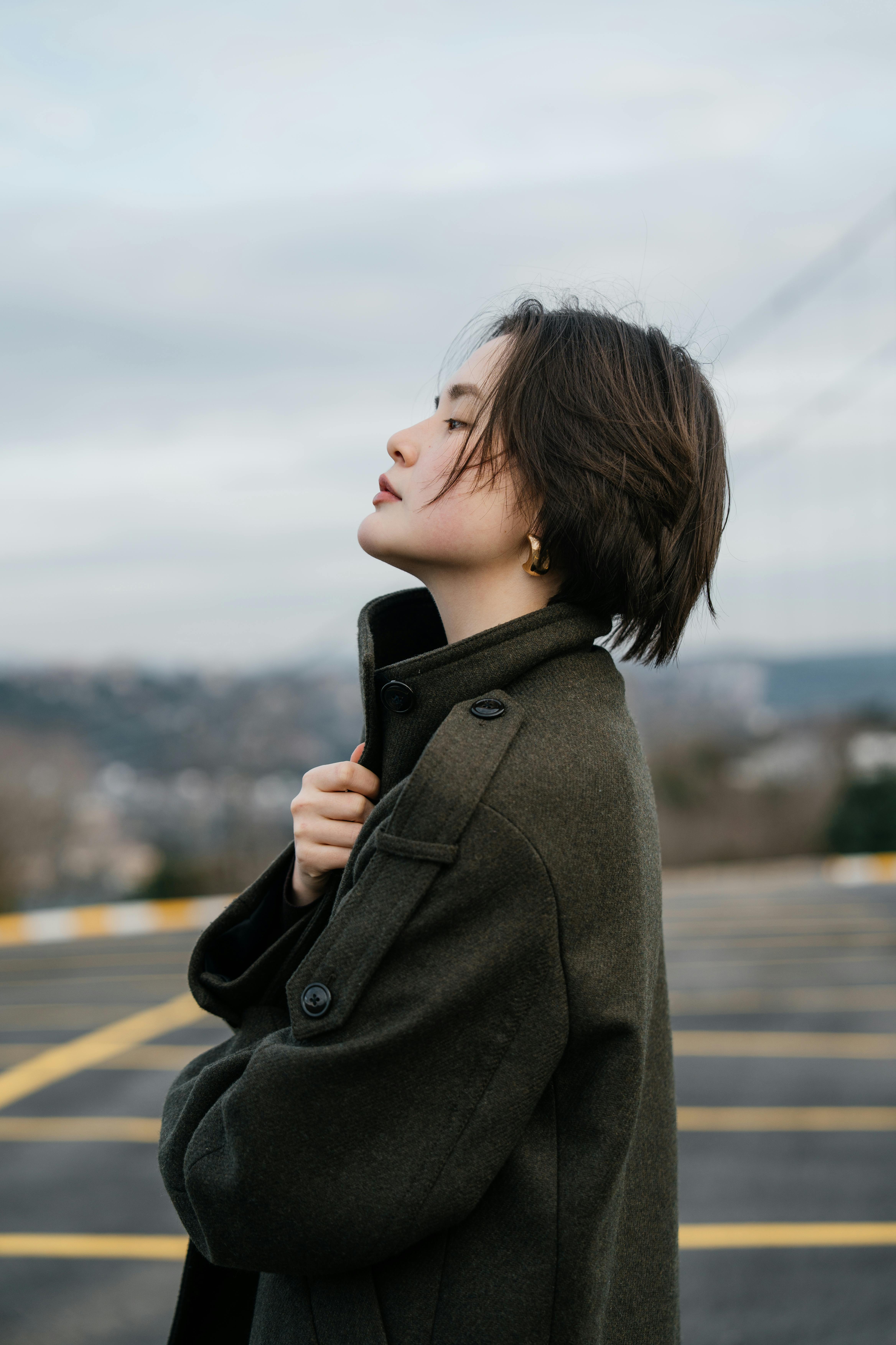 Side profile of a woman in a coat outdoors, looking pensive against a cloudy sky.