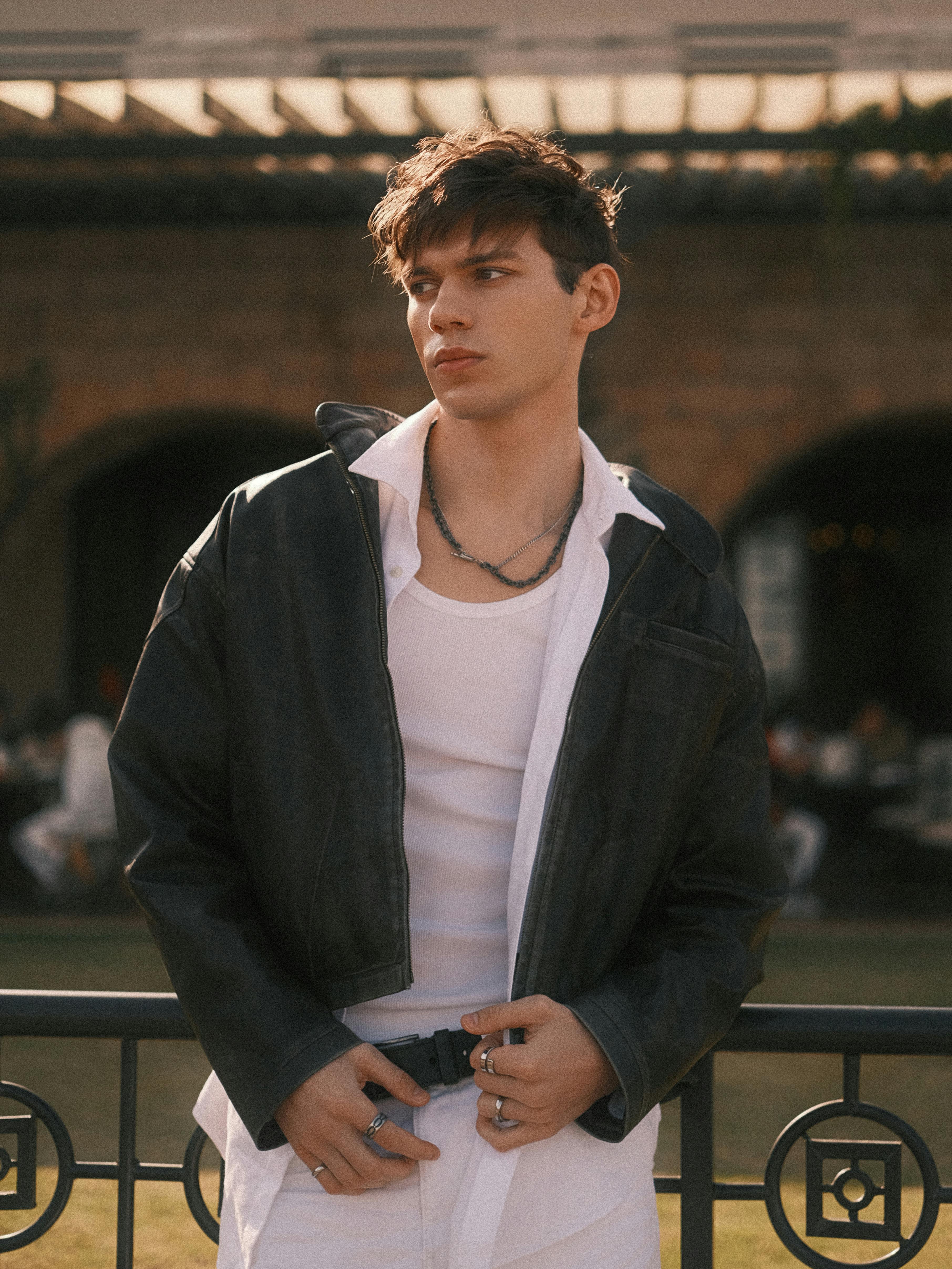 Stylish young man wearing leather jacket, posing casually outdoors in an urban setting.
