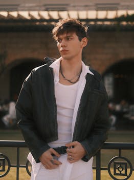 Stylish young man wearing leather jacket, posing casually outdoors in an urban setting.