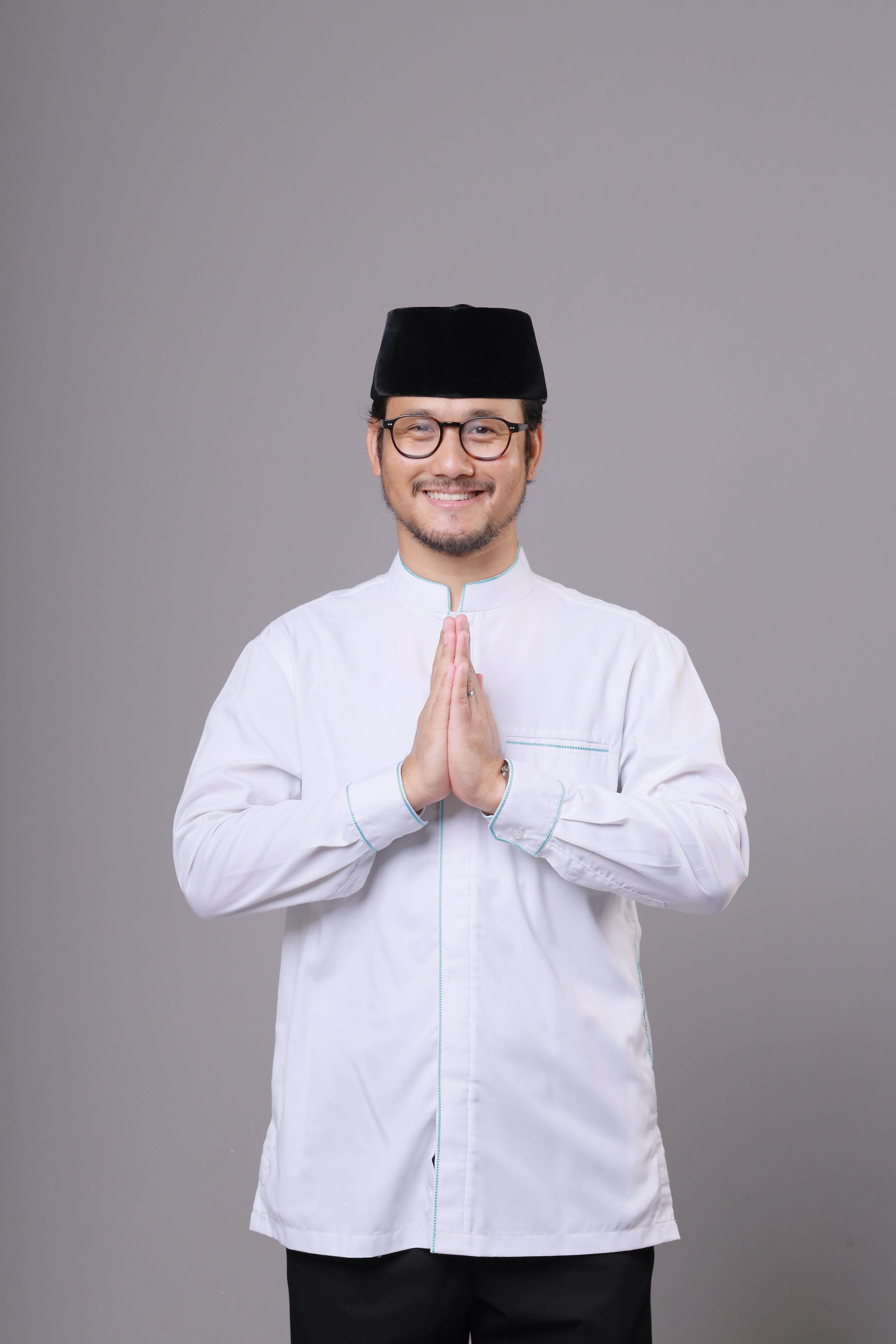 A man in traditional clothing with folded hands smiling against a gray background.