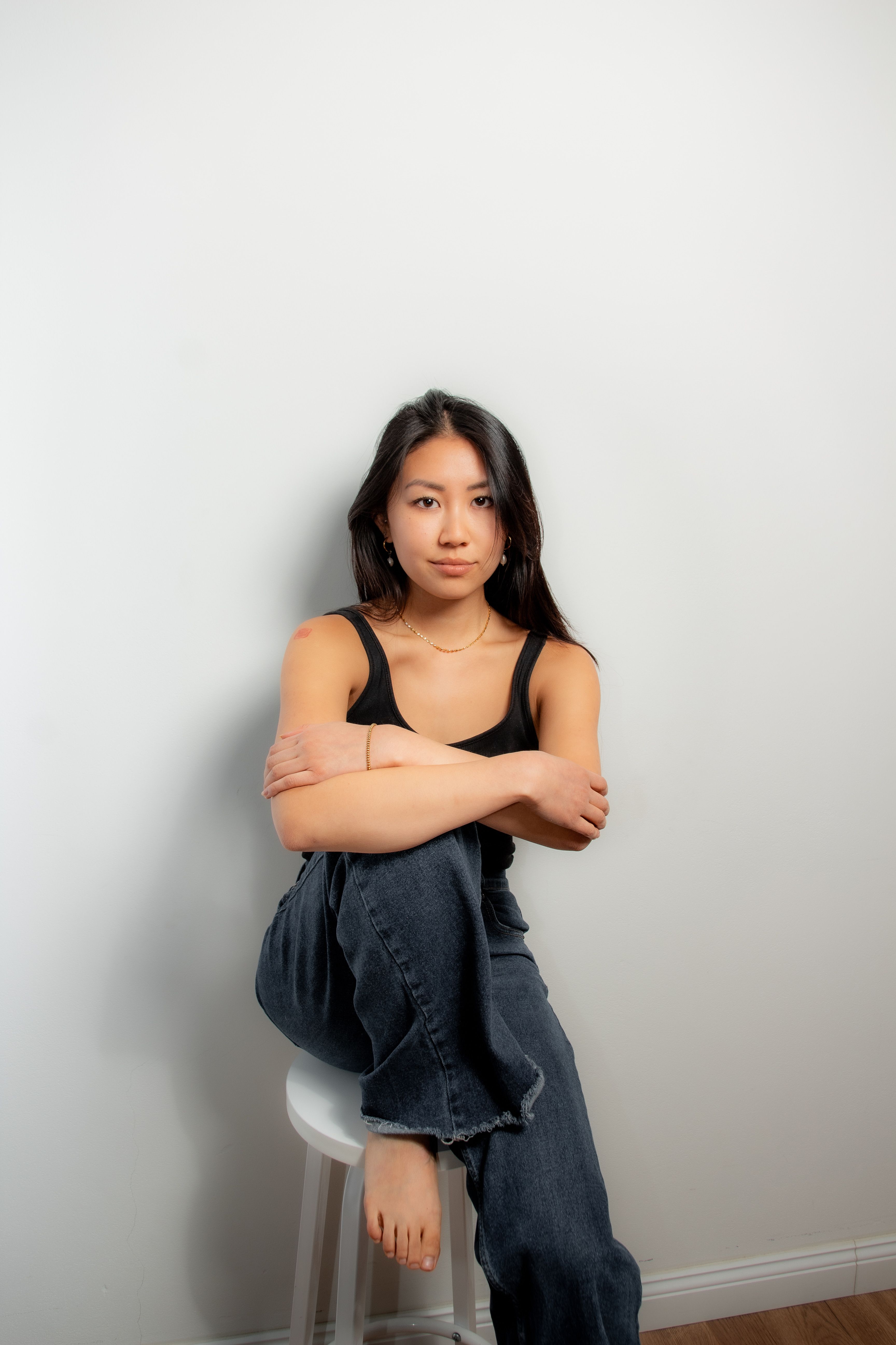 Portrait of an adult woman sitting casually on a stool indoors against a plain wall.