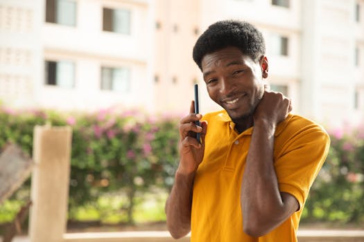 A young man in a yellow shirt smiling while talking on the phone outdoors.