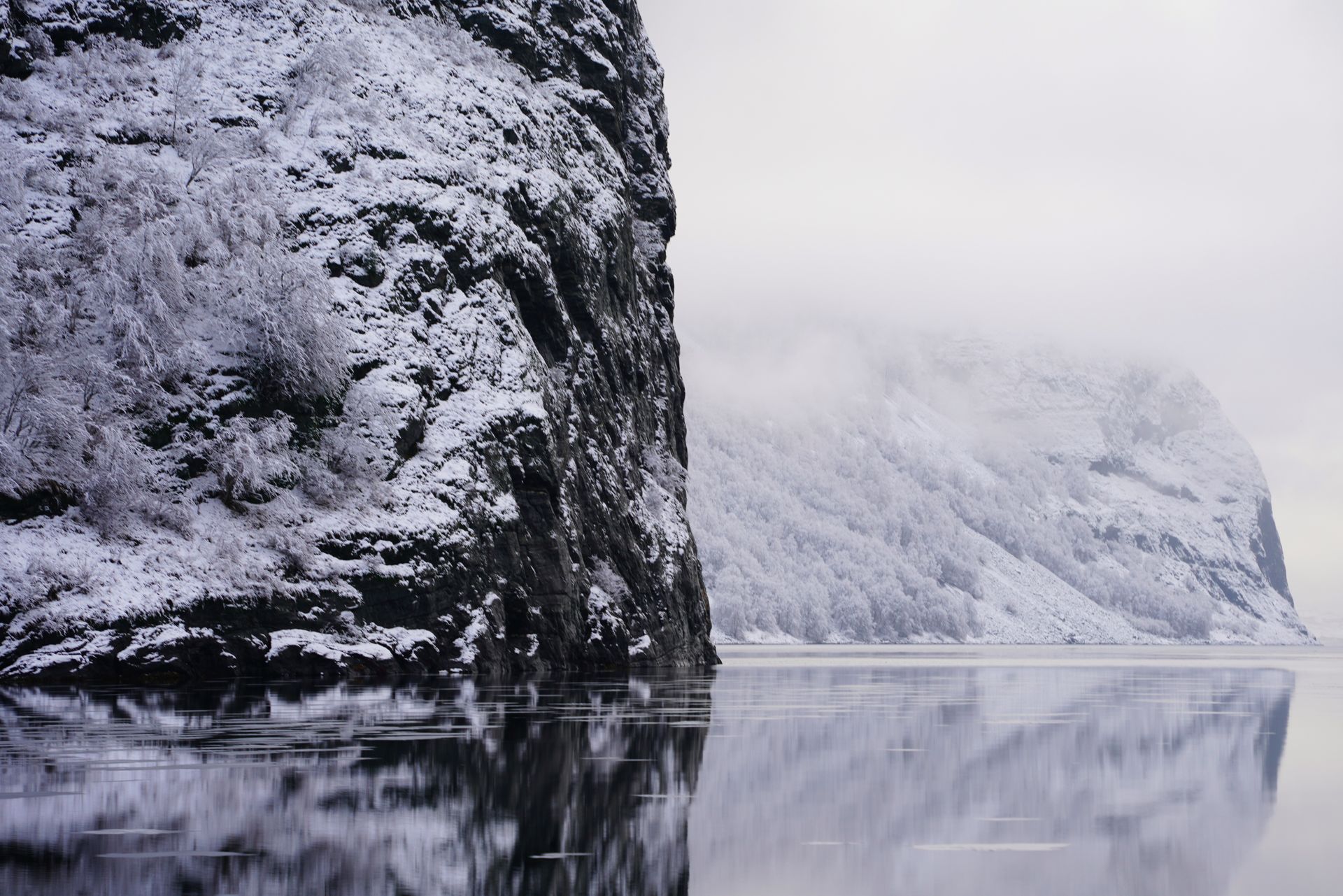 Frozen Norwegian fjord — snow-covered mountains reflected in dark icy water, frost on rocks, heavy storm clouds, cold atmosphere