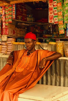 Man in traditional African clothing seated in a colorful market surrounded by packaged goods.