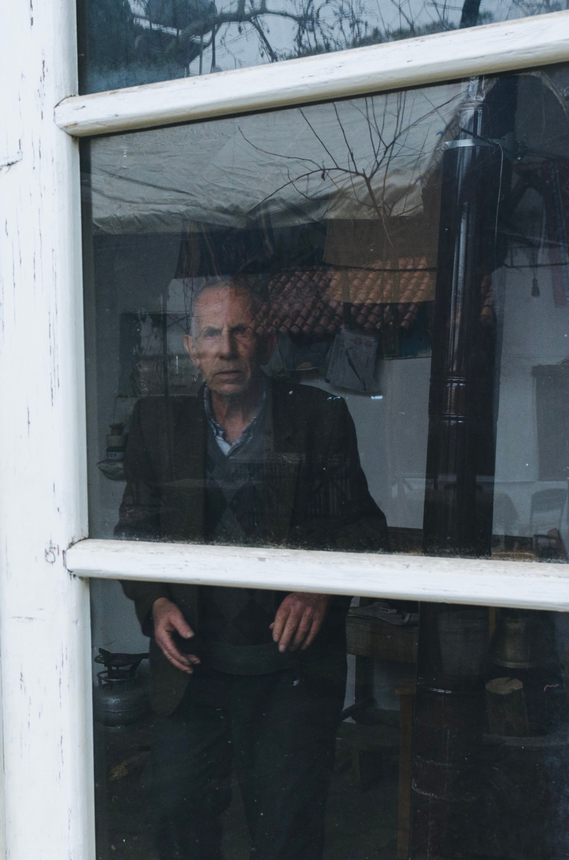 An elderly man gazes through a window from inside a rustic home.