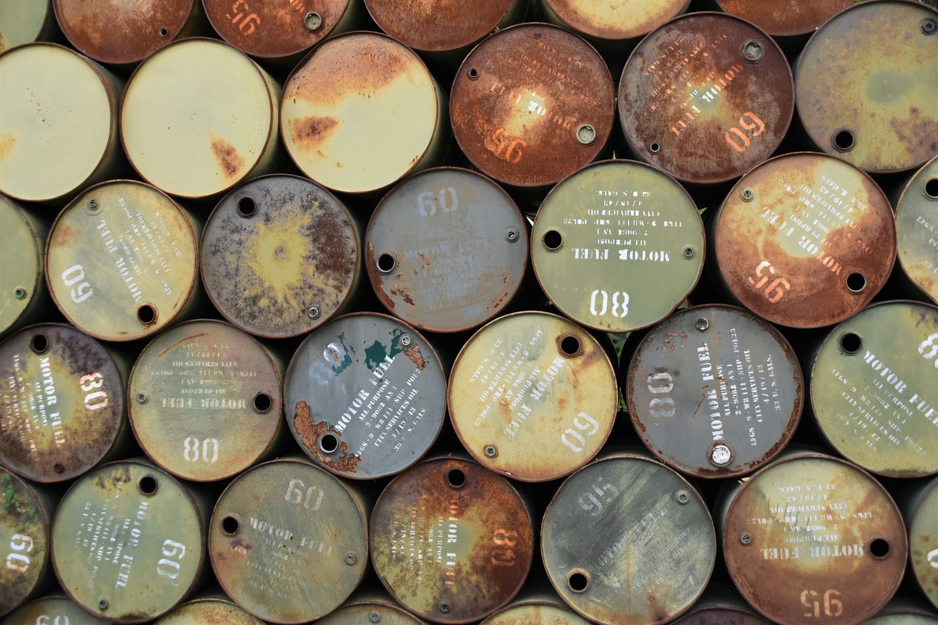 Pile of old, rusty oil barrels stacked outdoors with visible labels and numbers.