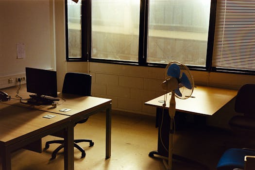 Quiet empty office with a desk, computer, and fan under warm afternoon sunlight.