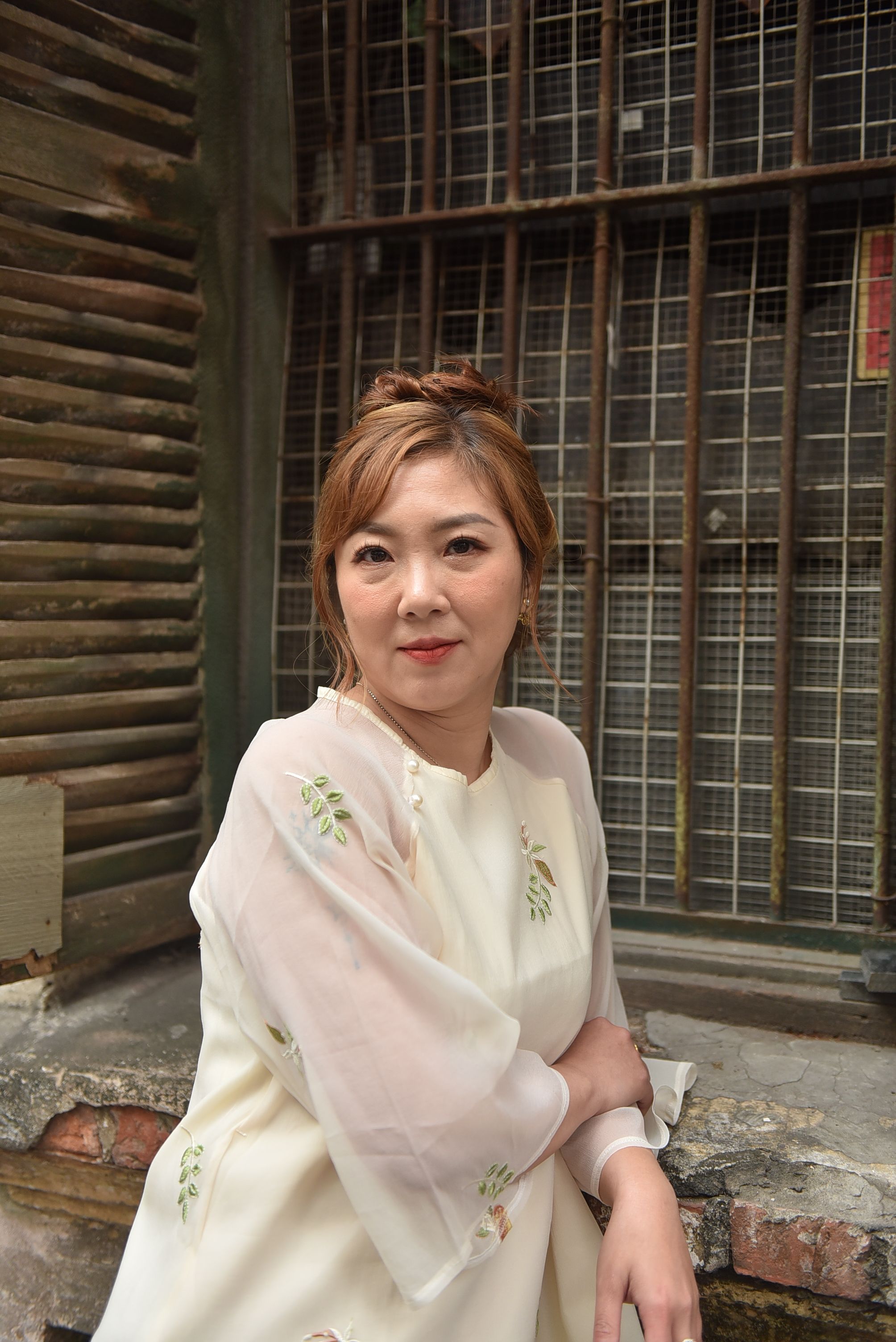 Charming woman in a white dress sits by a rustic urban backdrop, exuding elegance and warmth.