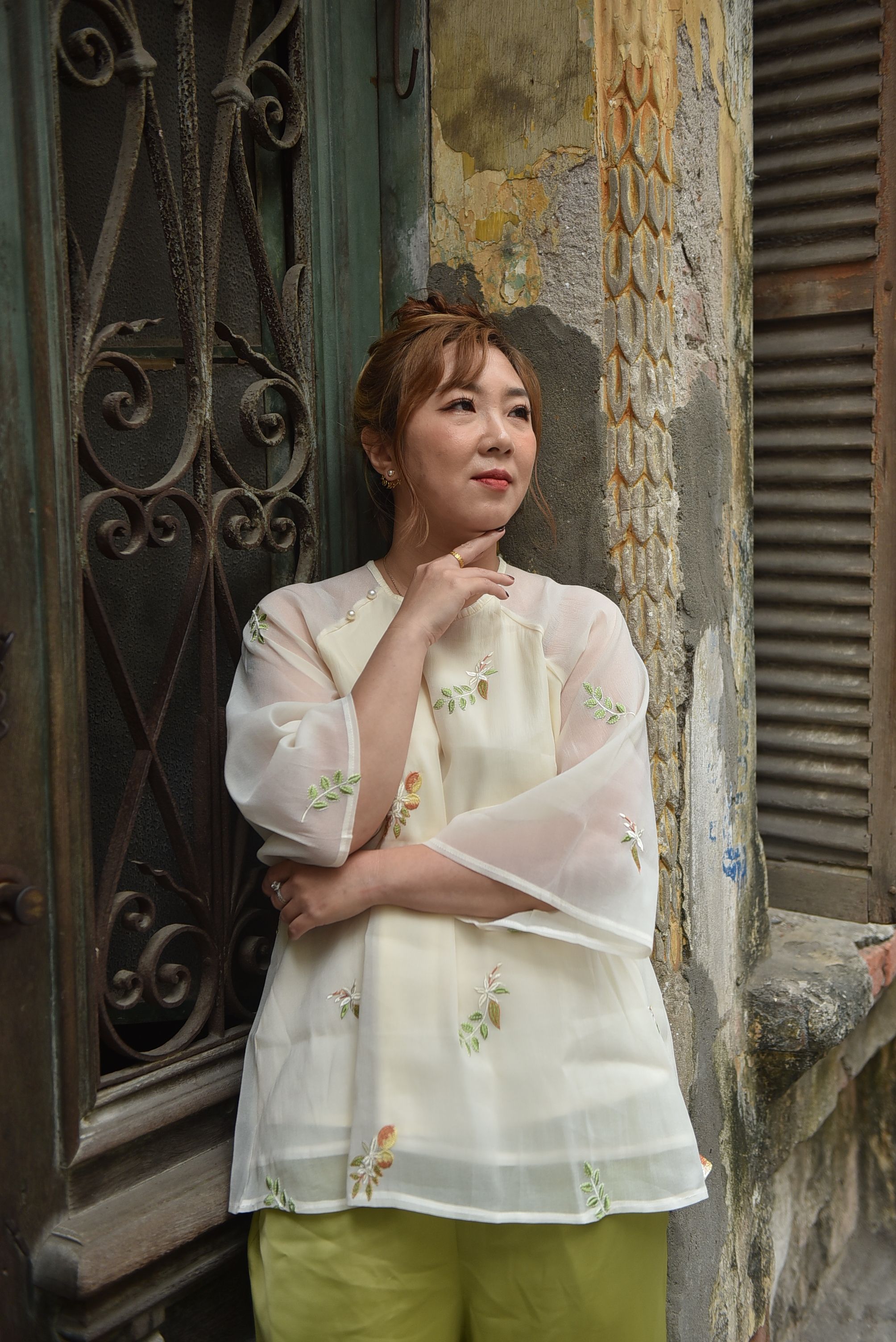 Asian woman in a floral blouse leans against a historic, textured wall.