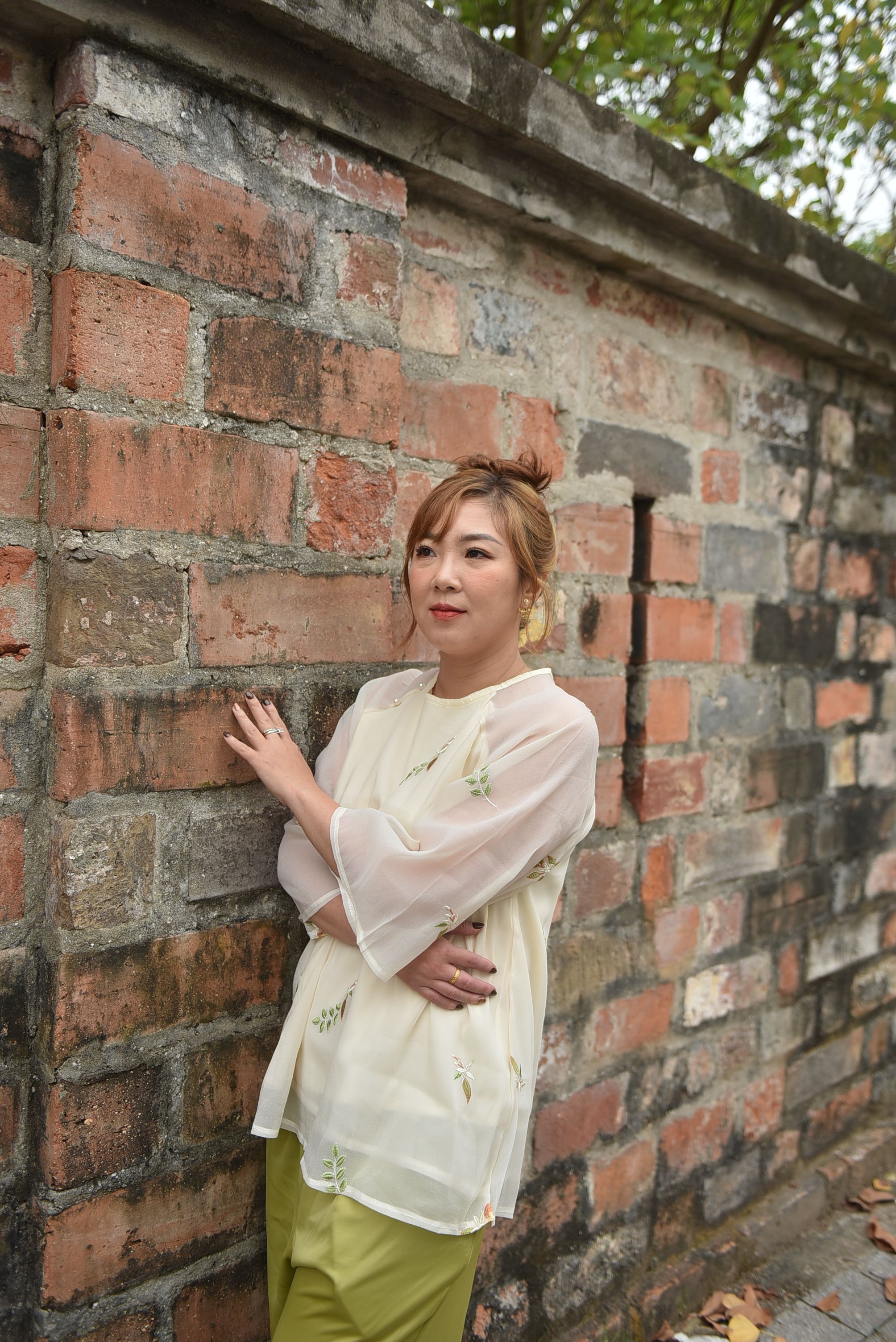 Elegant woman in light blouse poses by a rustic brick wall on a calm day.
