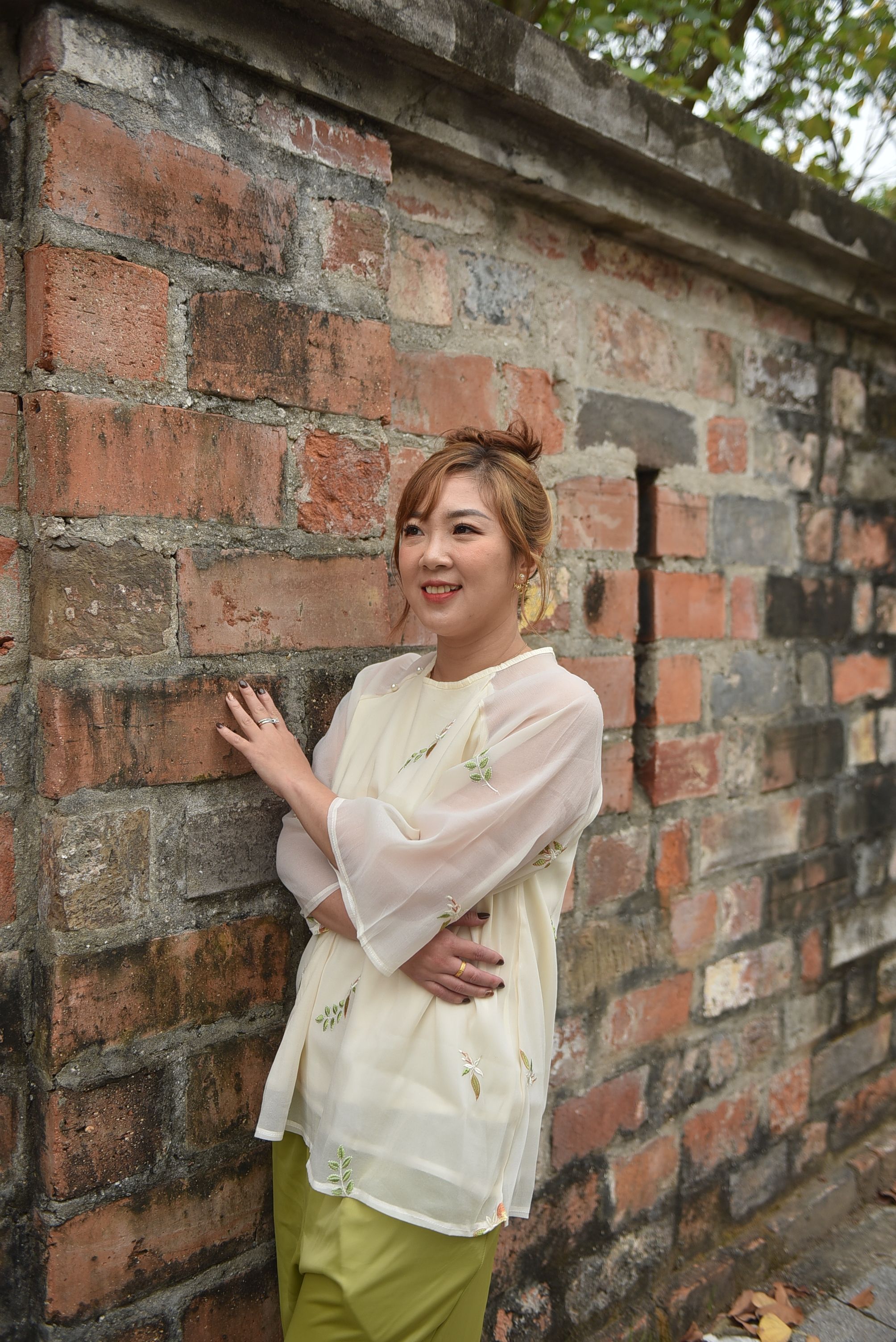 A cheerful woman in a white blouse stands against a vintage brick wall outdoors.