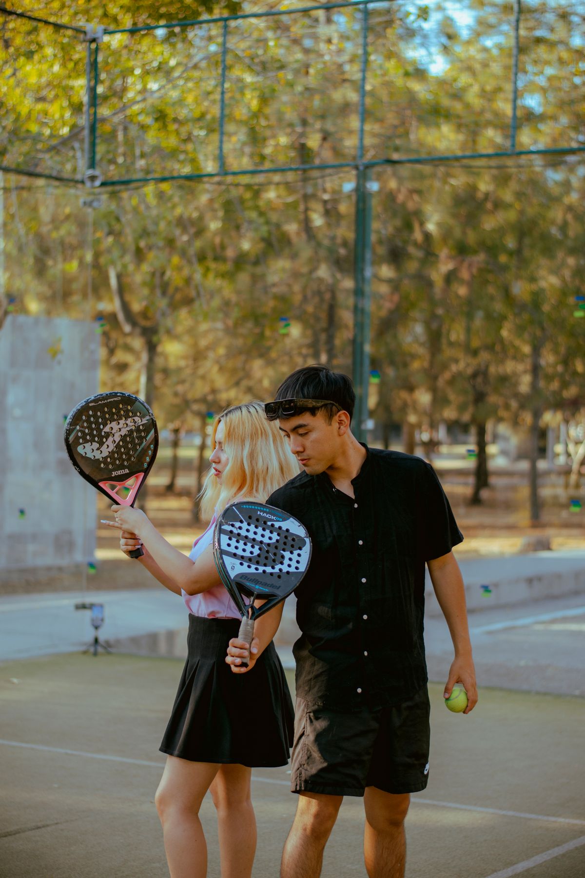 A coach shows a beginner how to hold her padel racket on an outdoor court under trees