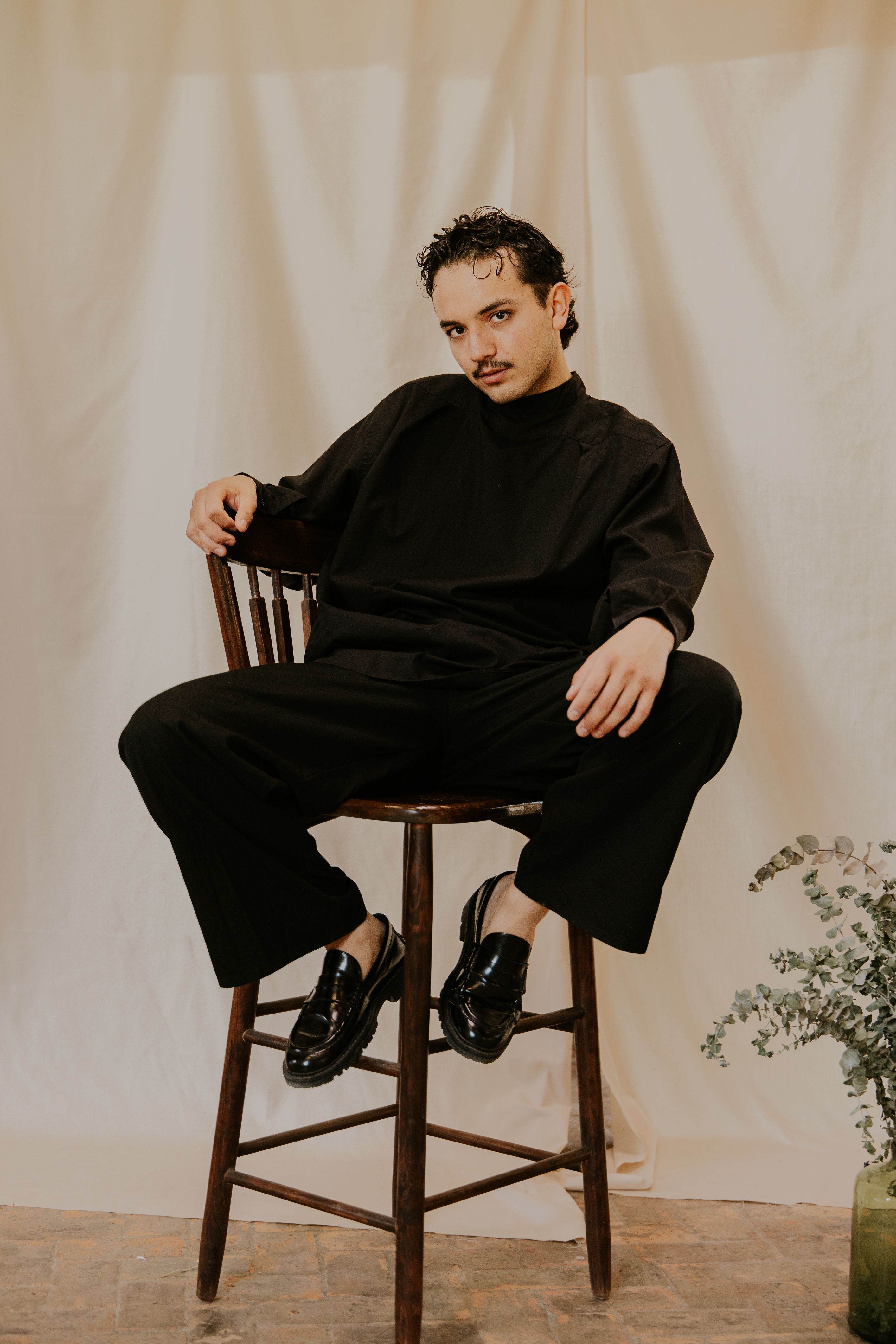 Elegant man in black attire poses confidently on a wooden chair, exuding modern fashion.
