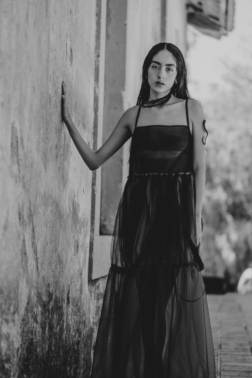 Free Black and white portrait of a young woman in a flowing black dress leaning against a wall outdoors. Stock Photo