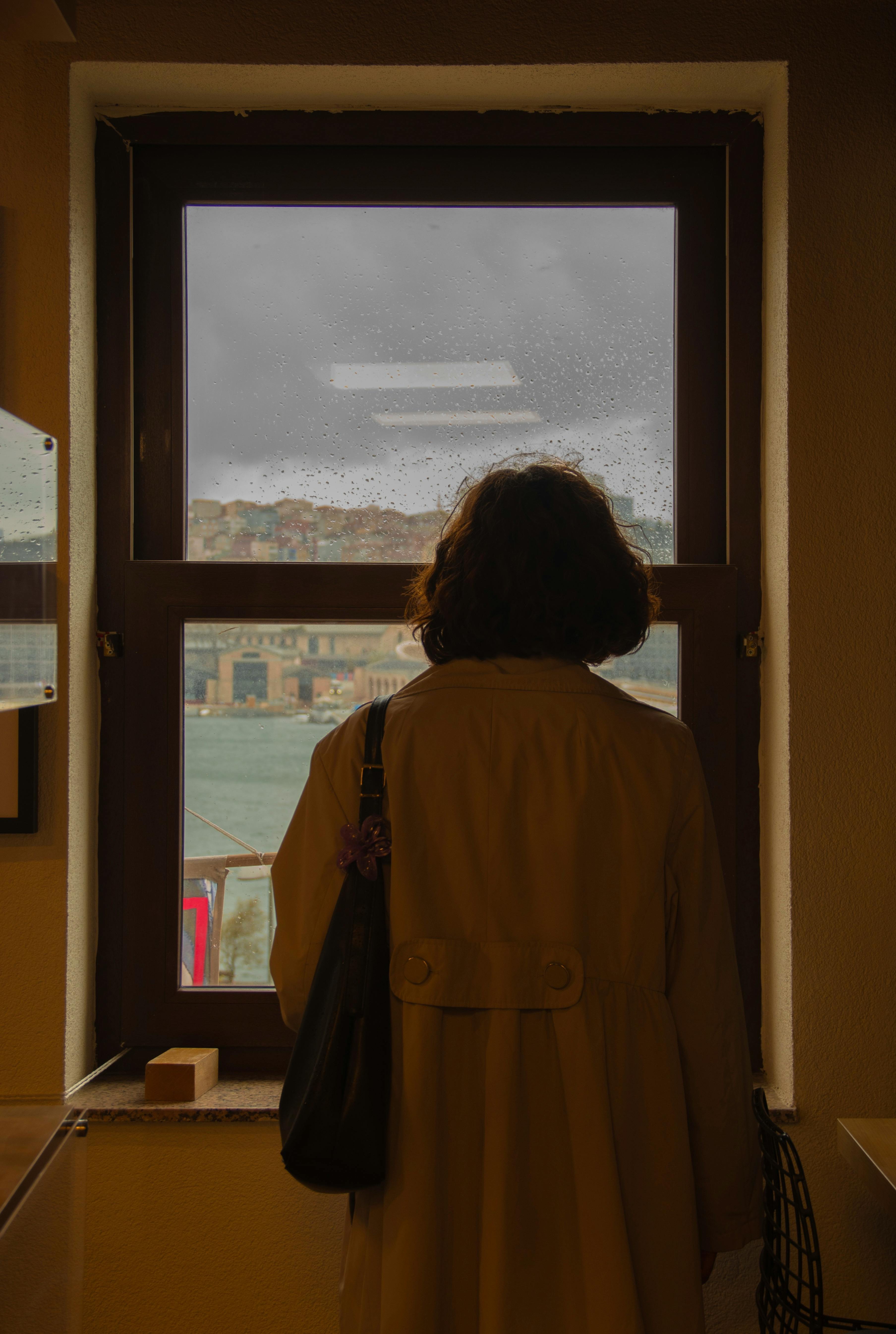 A woman gazes out a window at a rainy seascape, creating a contemplative mood.