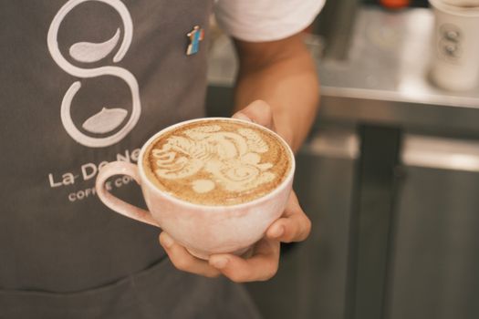 Close-up of a barista holding a cappuccino with intricate latte art, indoors in a coffee shop.
