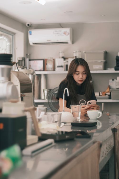 Free A young woman barista uses her smartphone at a cozy coffee shop counter. Stock Photo