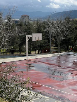 A wet basketball court surrounded by greenery and mountains in Isparta, Türkiye, on a cloudy day.