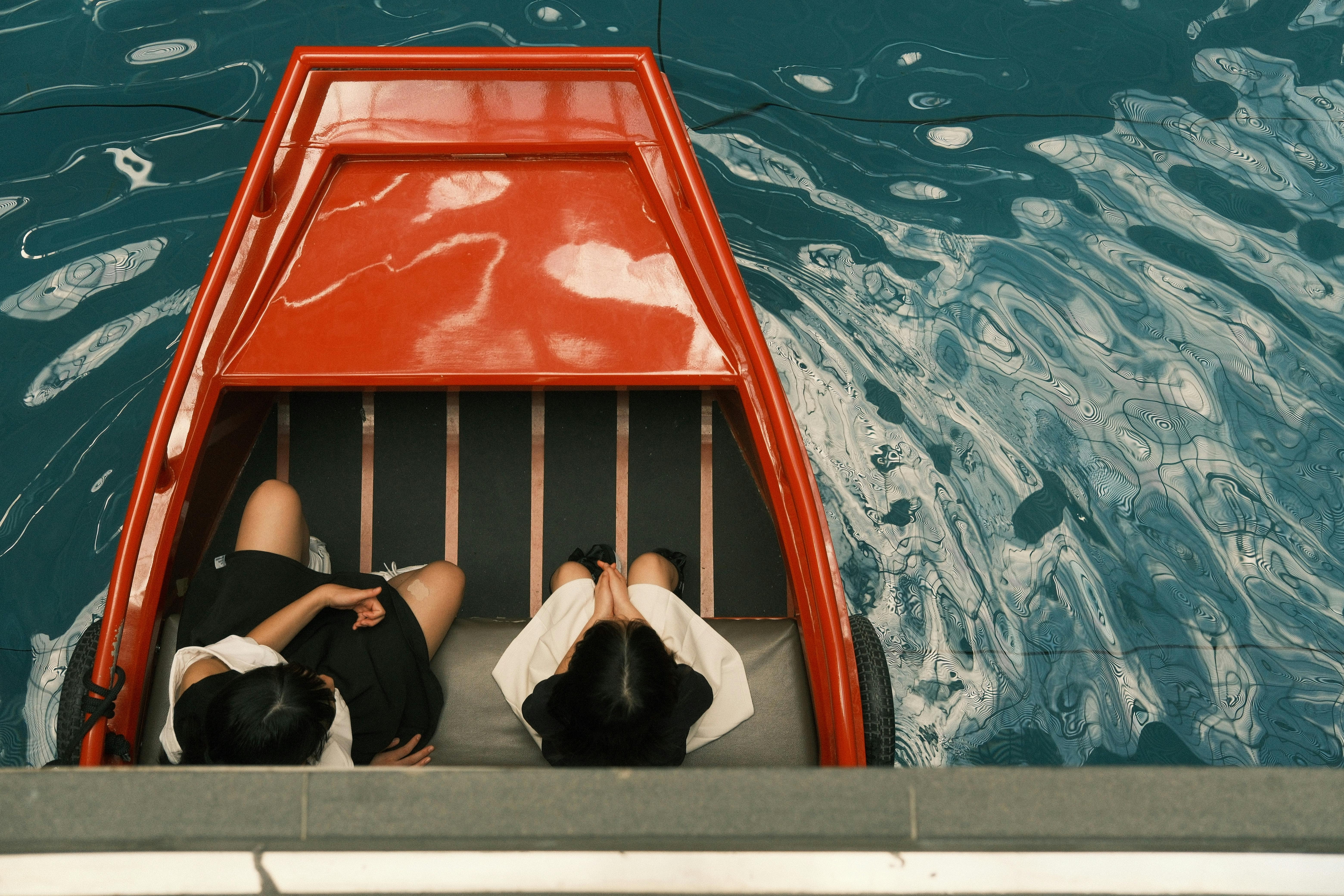 Two people sit in a red boat on vibrant blue water, seen from above.