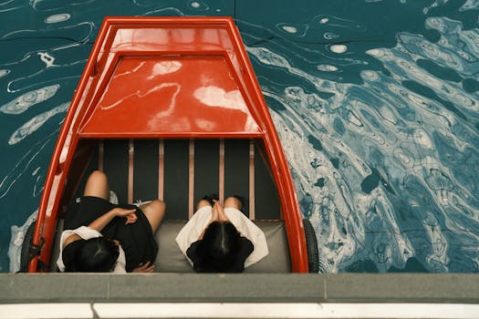 Two people sit in a red boat on vibrant blue water, seen from above.