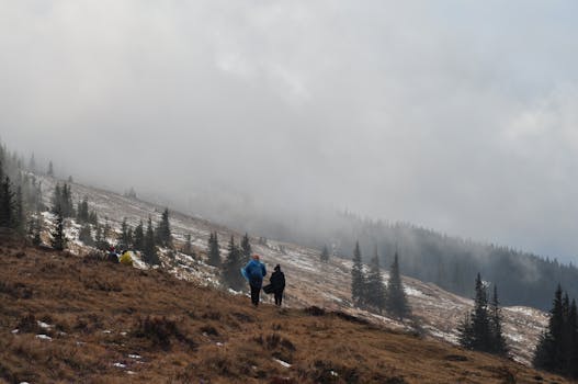 Two hikers traverse a foggy mountain landscape with sparse snow.