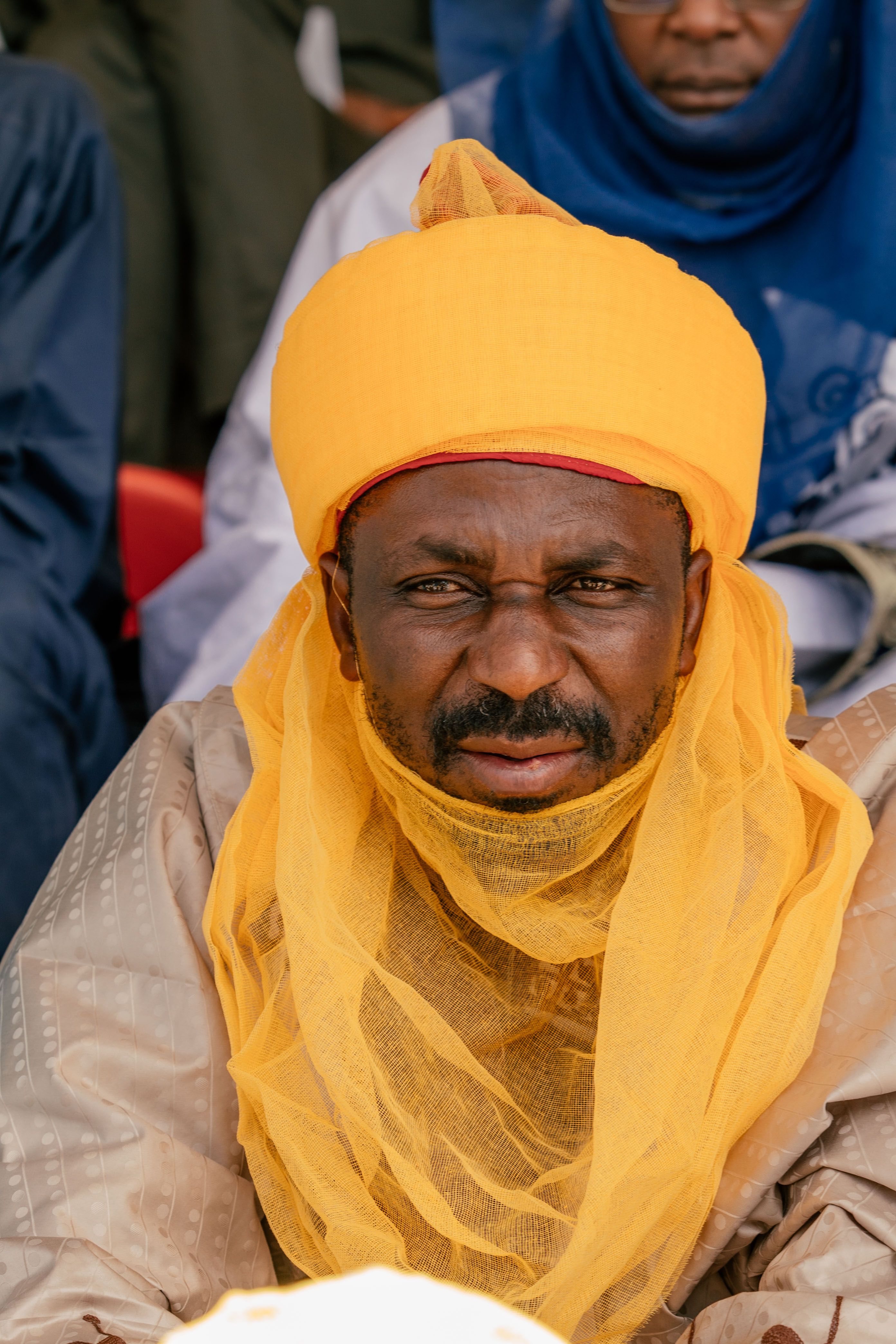 Close-up portrait of a man wearing vibrant traditional African attire, including a yellow turban and veil.
