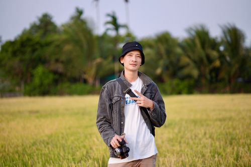 Free Smiling young man holding camera in a lush, green field under clear sky. Stock Photo