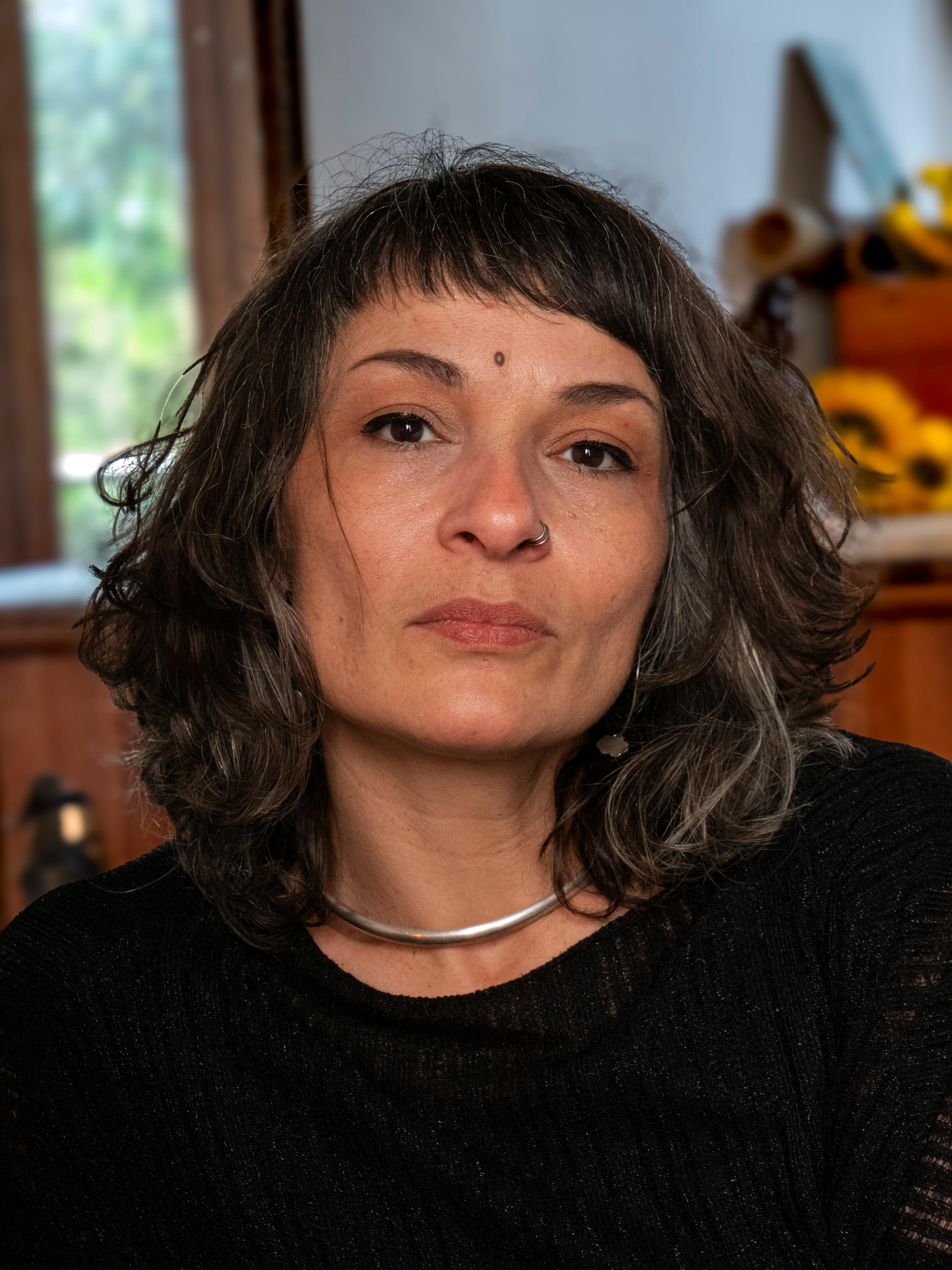 Close-up portrait of a woman with curly hair and nose ring indoors, looking thoughtful.