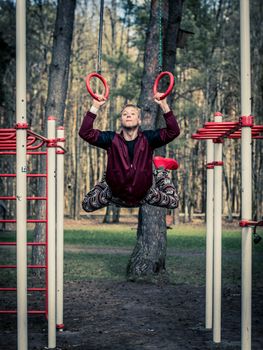 A young male gymnast executes an impressive move on outdoor rings in a Vilnius park, showcasing fitness and agility.
