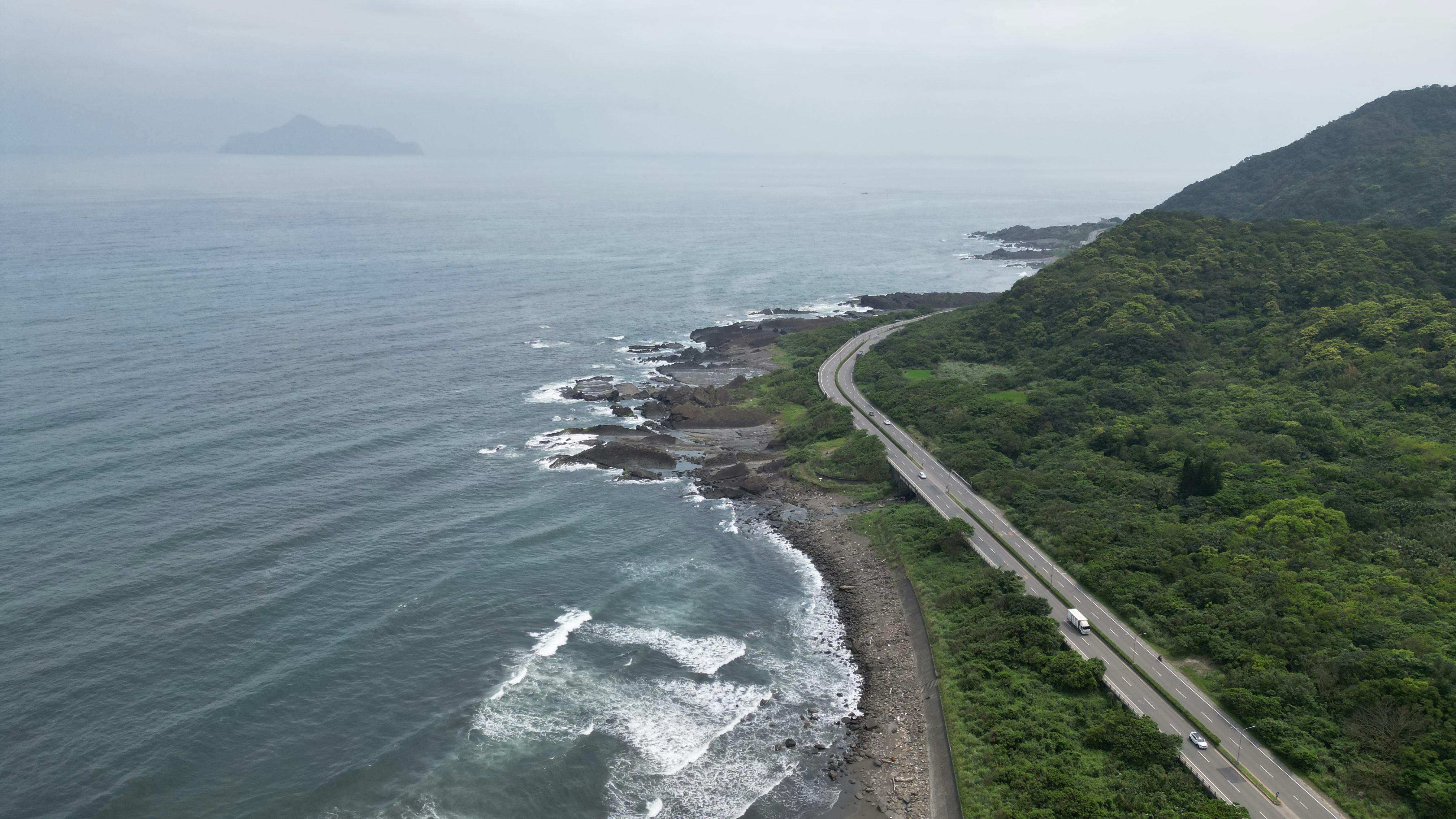 Aerial view of a picturesque coastal road winding along a lush green hillside next to the ocean waves.