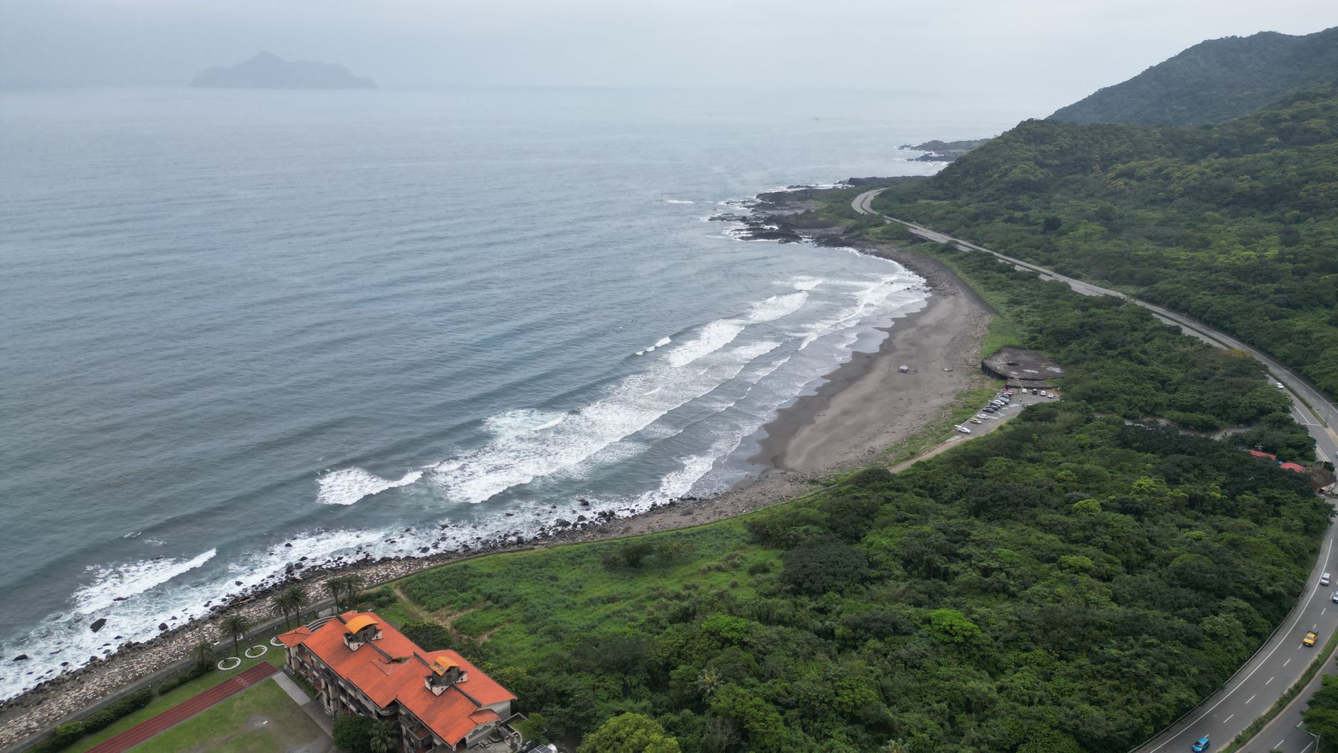 Aerial view of Taiwan's lush coastline with waves crashing on the shore, surrounded by greenery and a winding road.