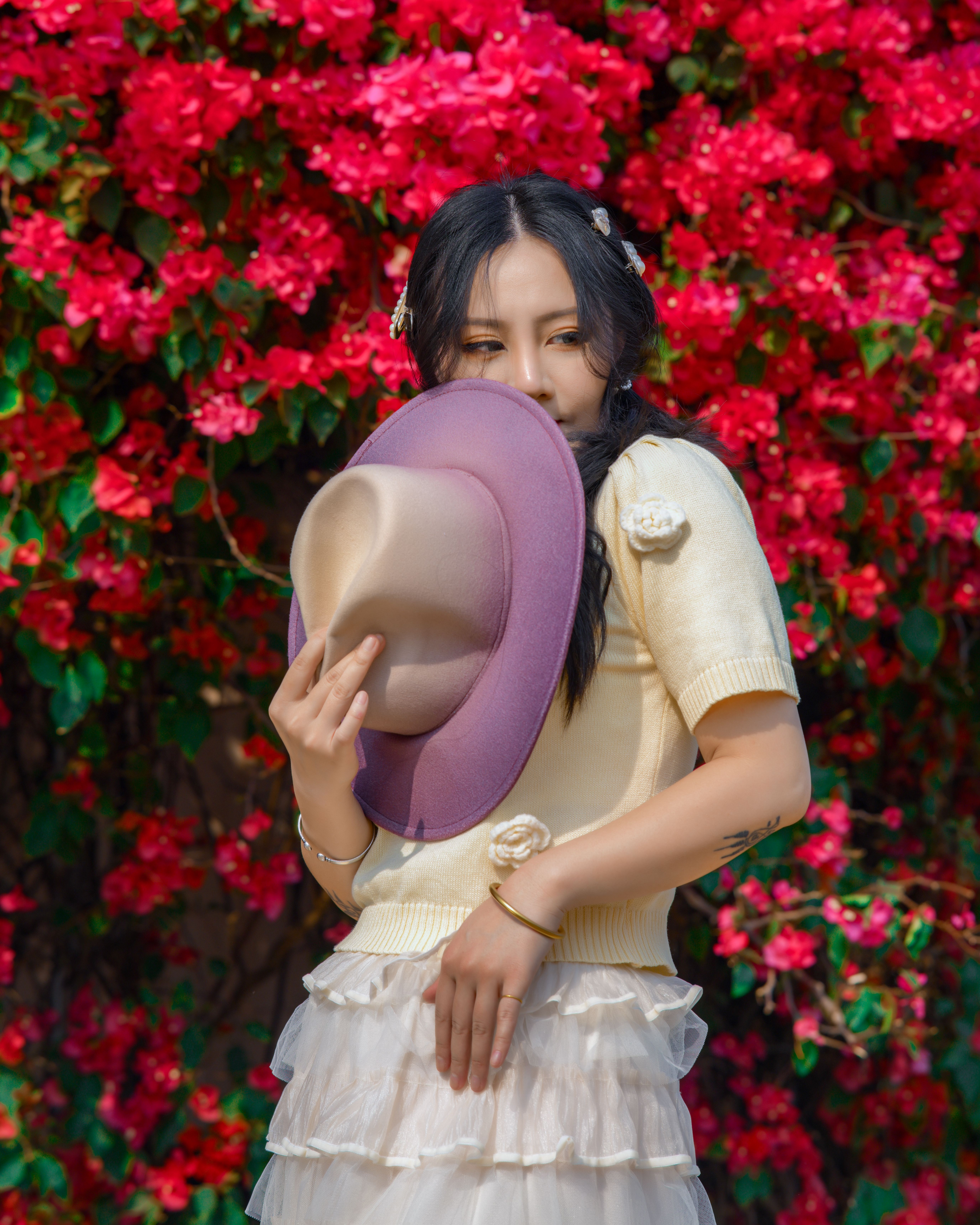 Woman posing with hat near vibrant pink bougainvillea hedge, exuding elegance.