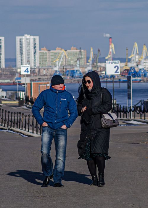 Free A couple strolls along a waterfront, bundled up in winter clothing on a sunny day. Stock Photo